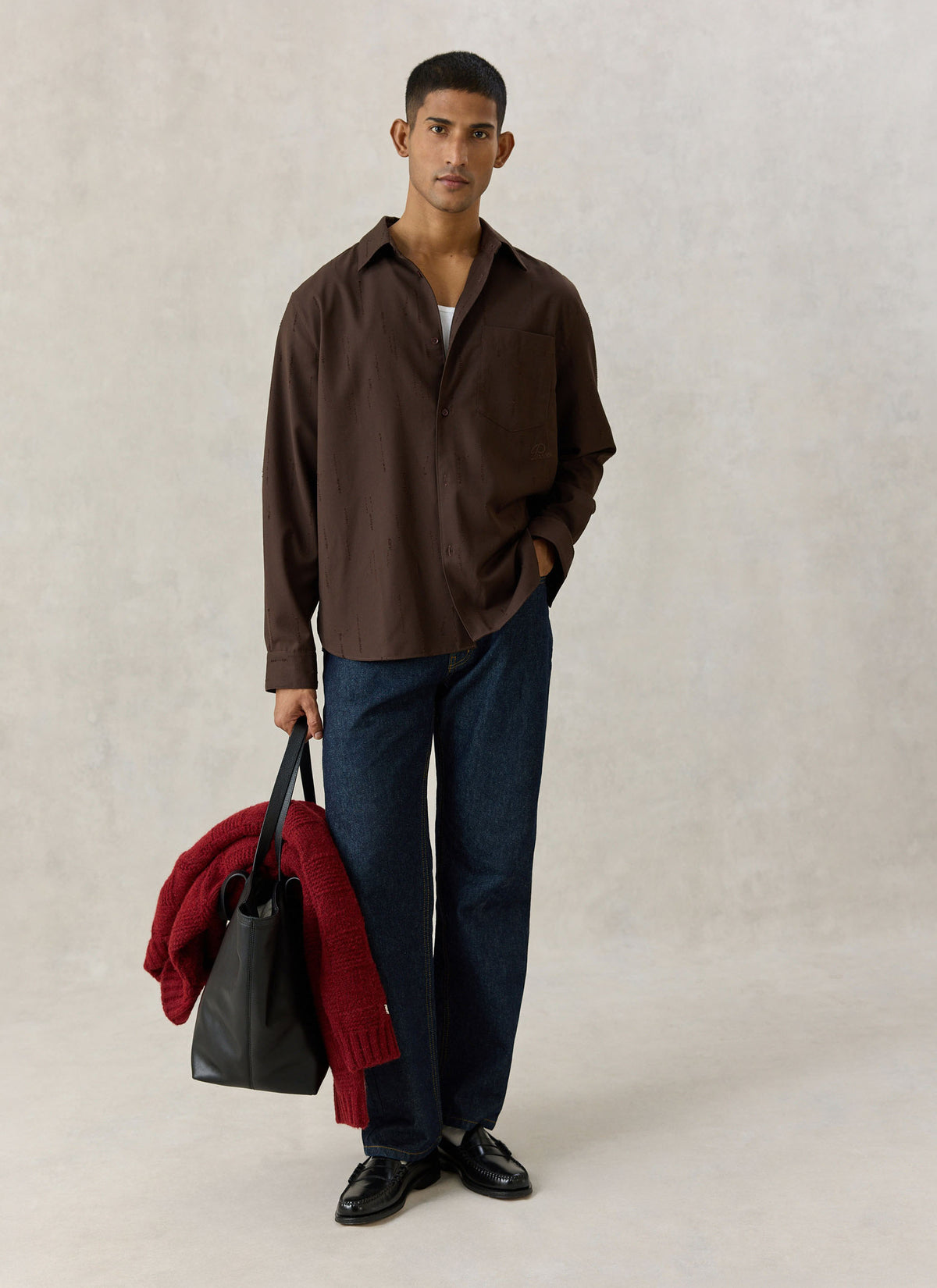 A model wearing the espresso brown Percival Rowley textured jacquard shirt with dark jeans and holding a bag and red knit.