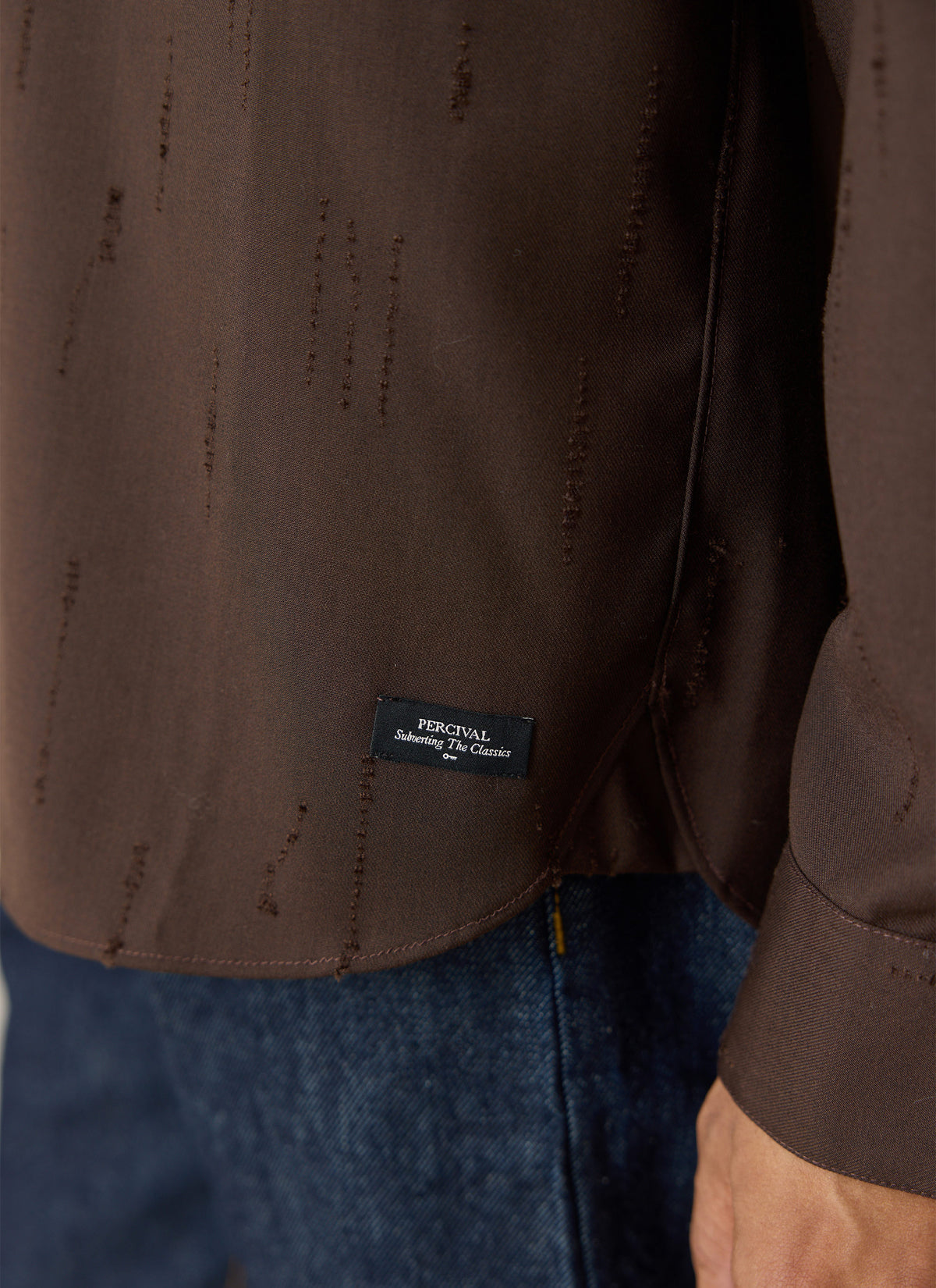 A close-up of the espresso brown Percival Rowley textured jacquard shirt showing the hem label and denim jeans.