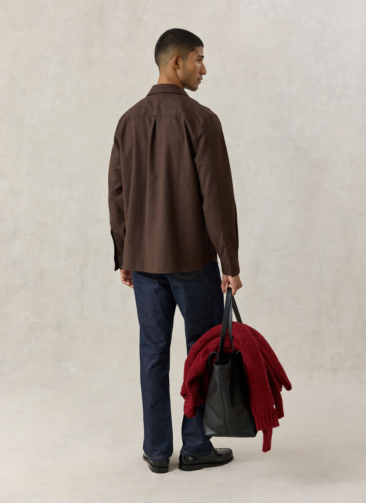 A model from the back wearing the Percival brown Rowley textured jacquard shirt with dark denim jeans and carrying a bag.