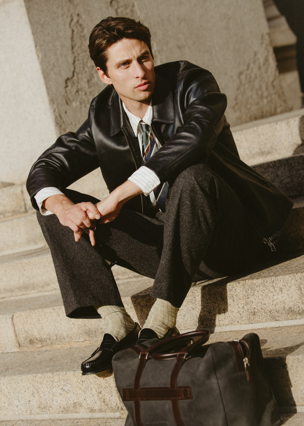 A model wearing Percival charcoal brushed-wool trousers and a leather jacket sitting outdoors on stone steps.