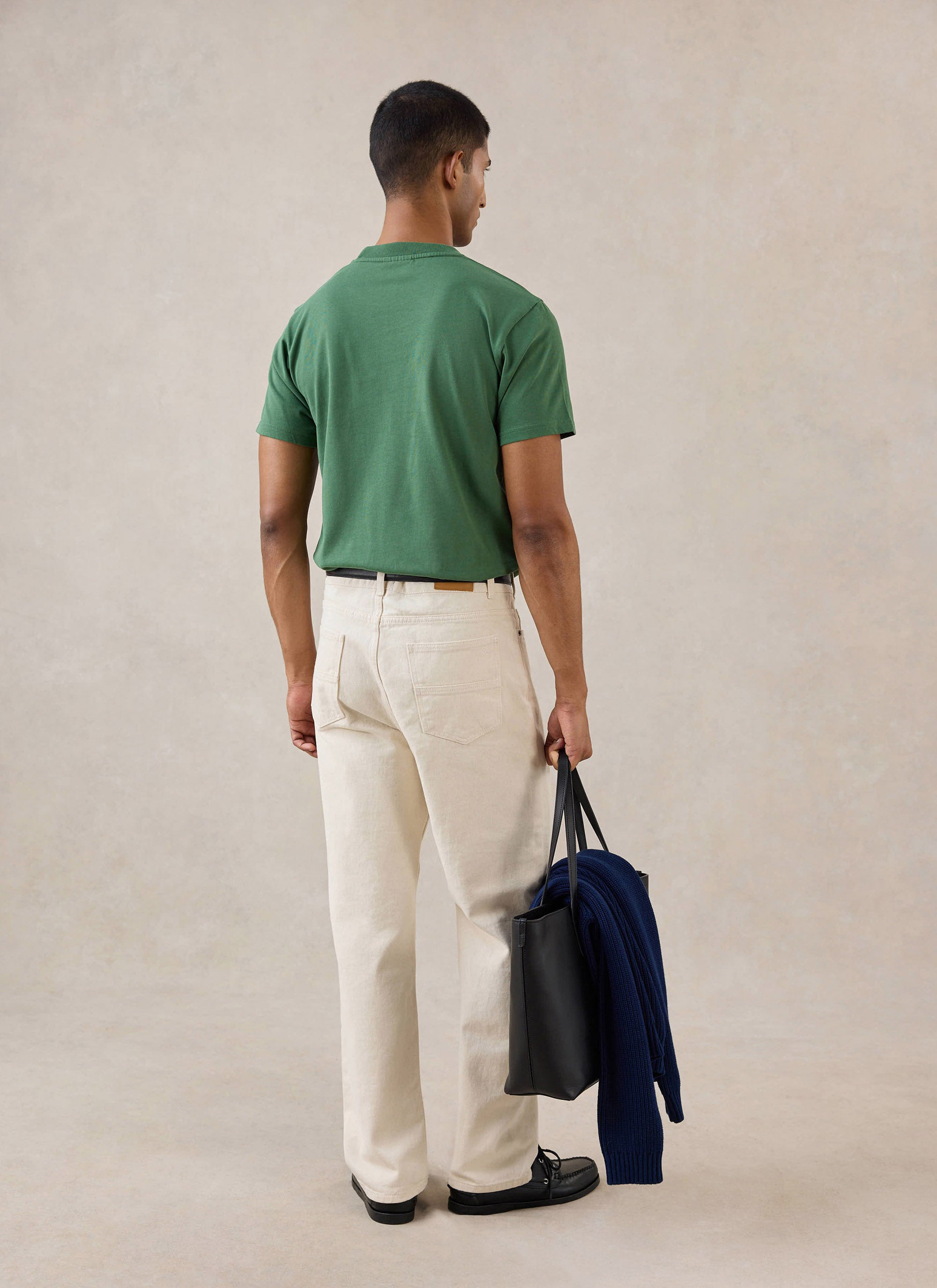 A rear view of a model wearing the forest Percival T-shirt, styled with cream trousers, black shoes, and holding a tote bag.