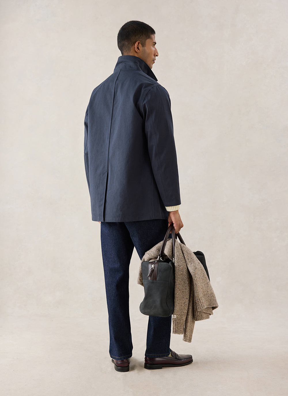 A model wears the navy Percival Sherlock short trench coat from behind, holding a dark bag and knitwear.