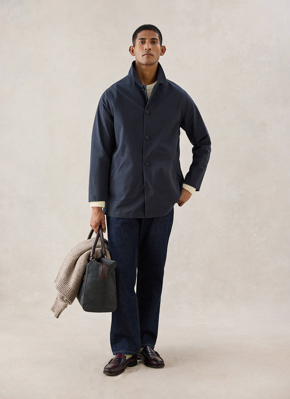 A model wearing the navy Percival Sherlock short trench coat, dark trousers, and holding a bag and jumper.