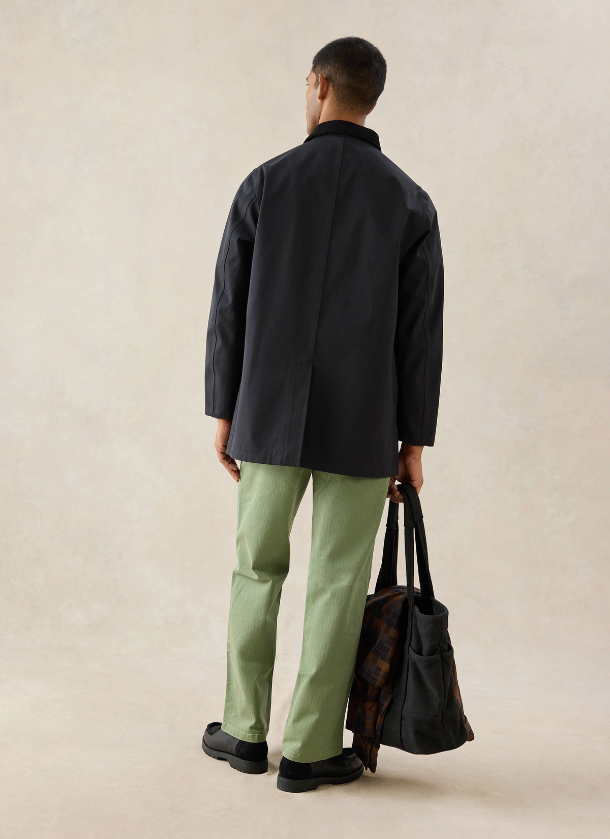 A model from behind wears the black Percival Sherlock Short Trench Coat, styled with green trousers and carrying a tote bag.