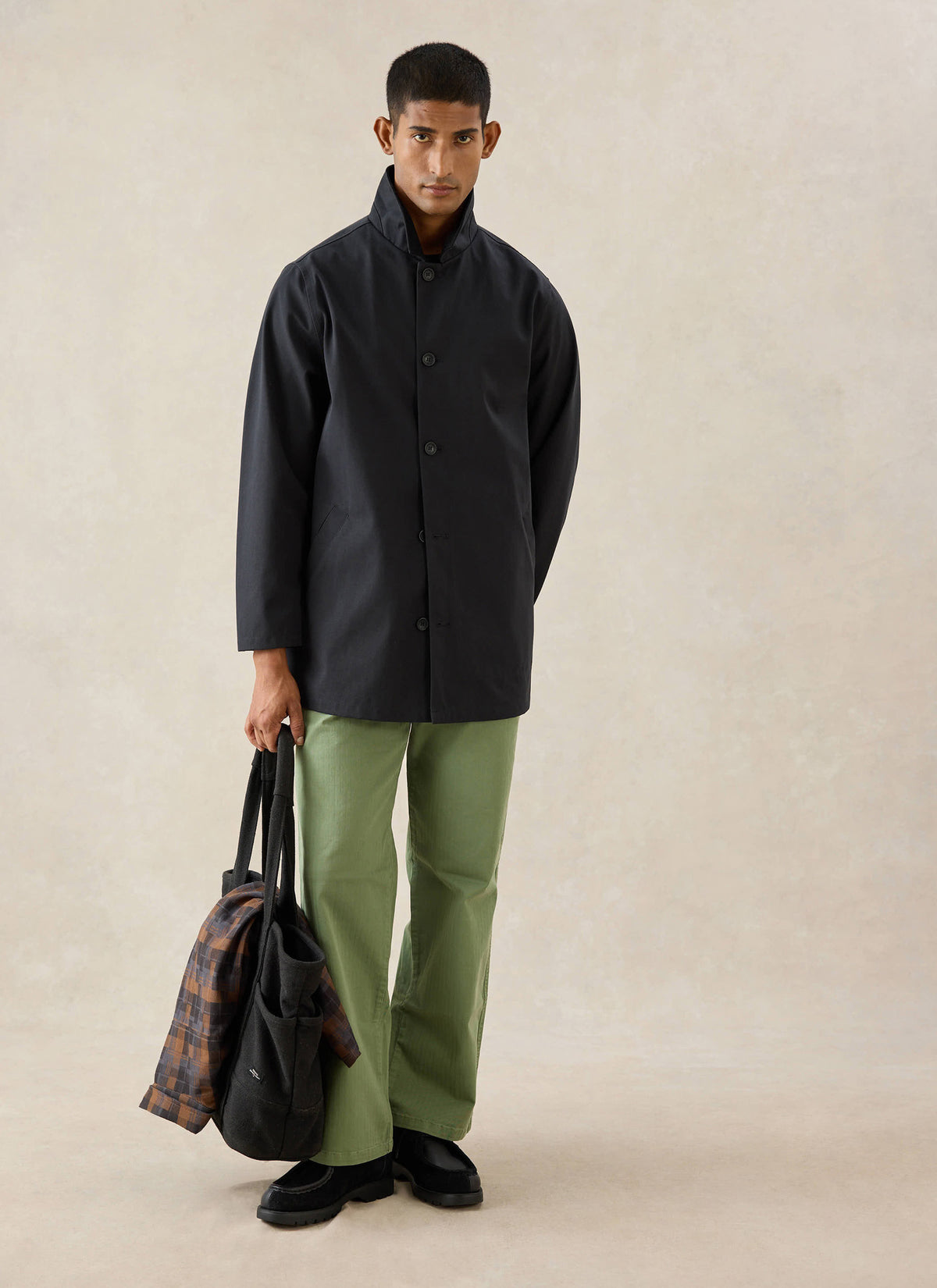 A model wears the black Percival Sherlock Short Trench Coat, styled with green trousers and carrying a bag in a studio shot.