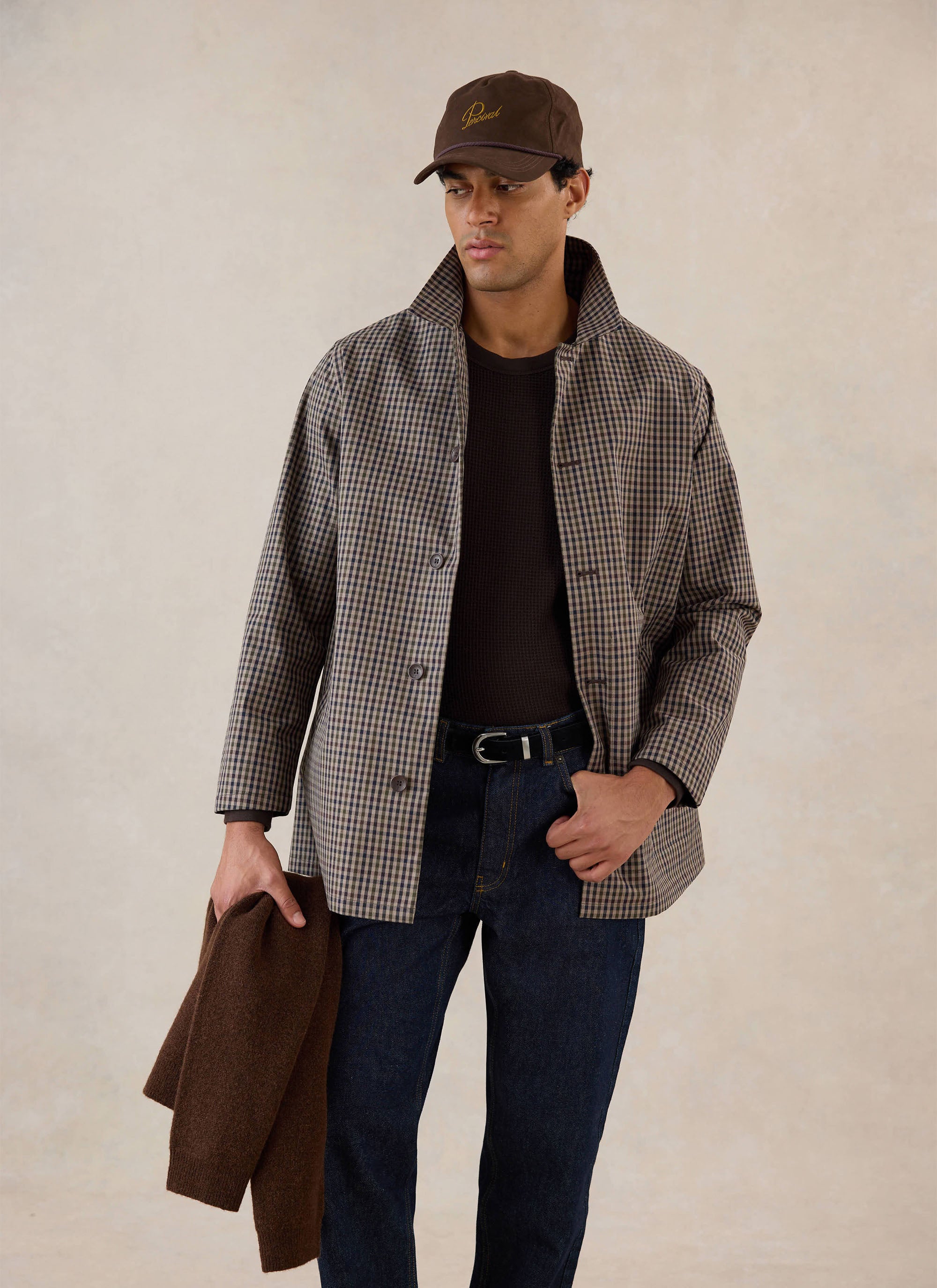 A model wears the Watcher Check Sherlock Short Trench Coat from Percival with dark jeans and a brown cap in a studio.