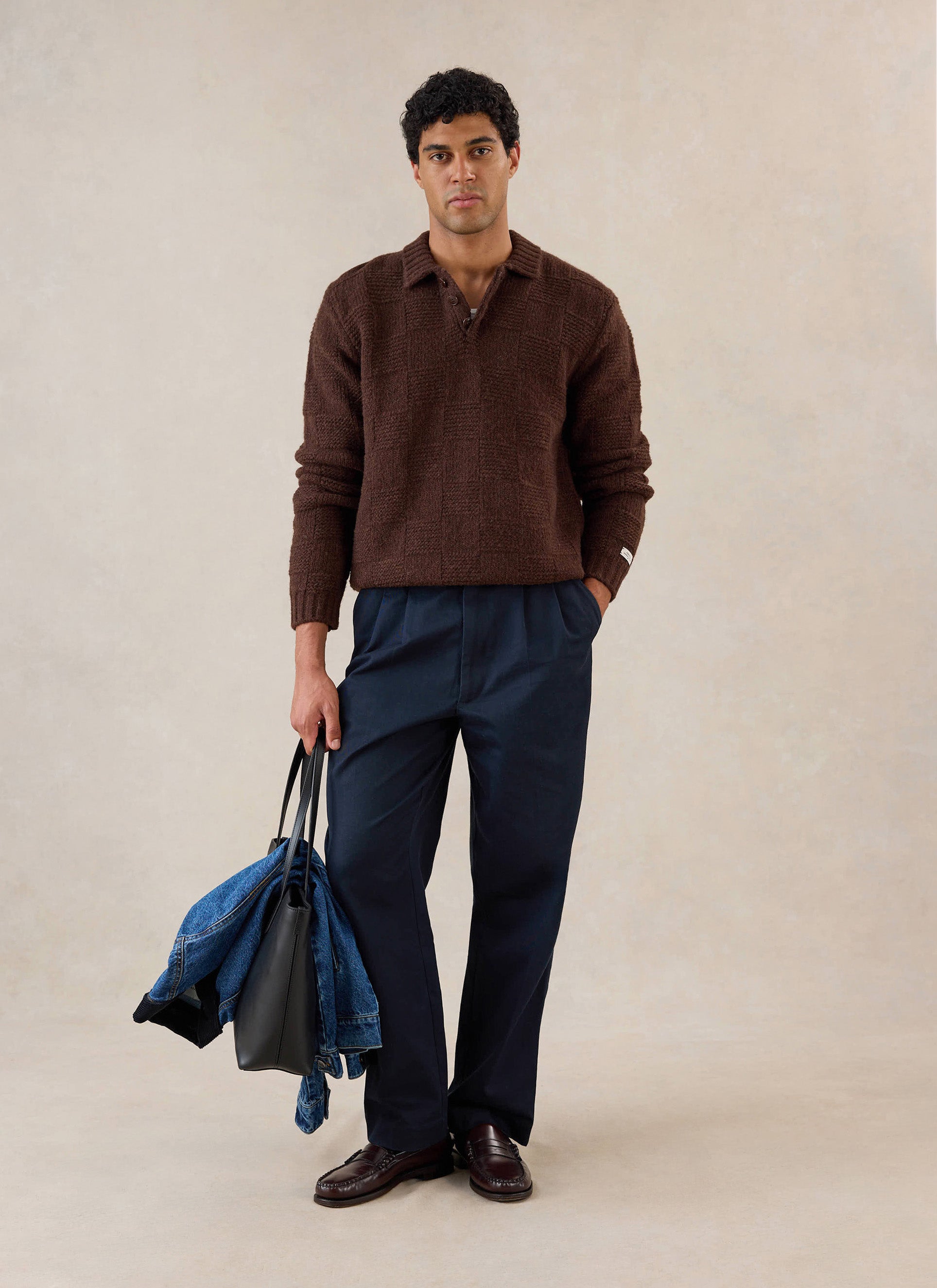 A model wears the brown Percival Shugart boxy knit jumper, styled with navy trousers, a black tote bag, and loafers.