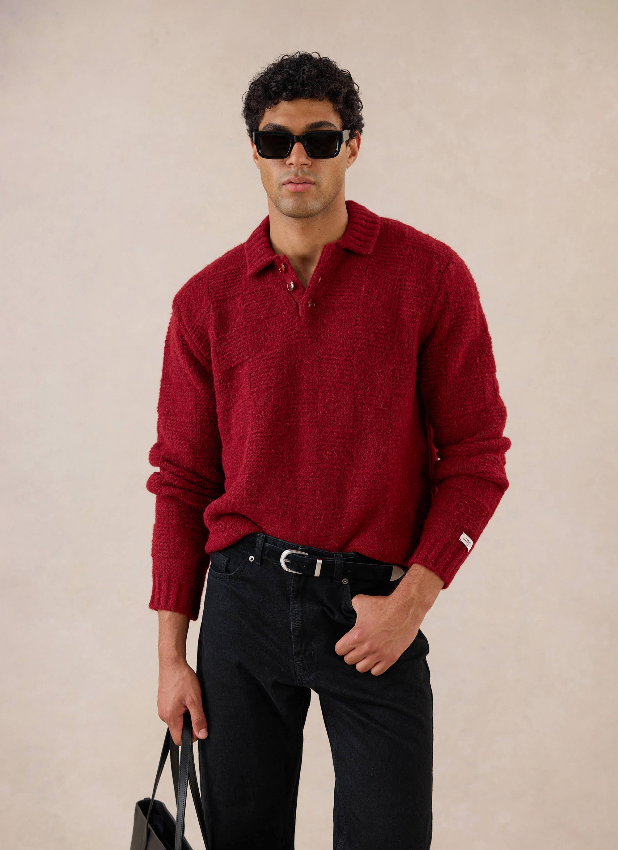 A model wears the merlot Percival Shugart knit jumper, styled with black jeans and sunglasses in a studio.
