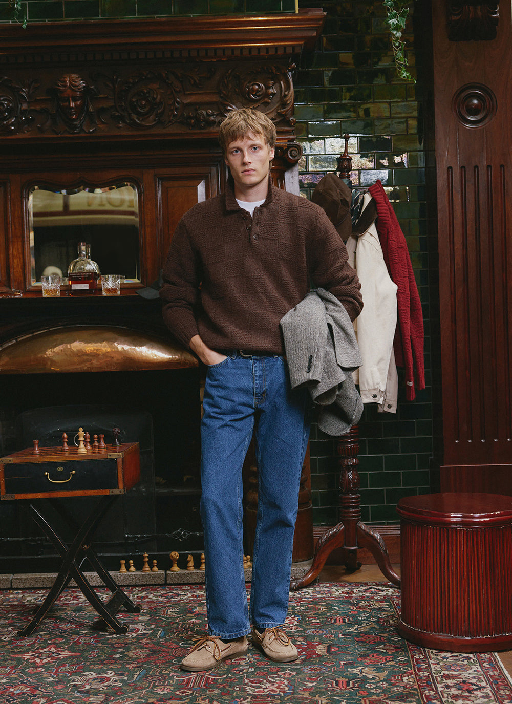 A model wears the brown Percival Shugart alpaca wool knit jumper with blue jeans in a pub interior setting.