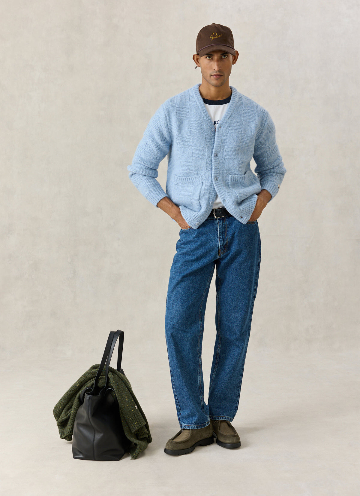 A model wearing the Percival light blue alpaca wool knit cardigan with jeans, a brown cap, and a black tote bag.
