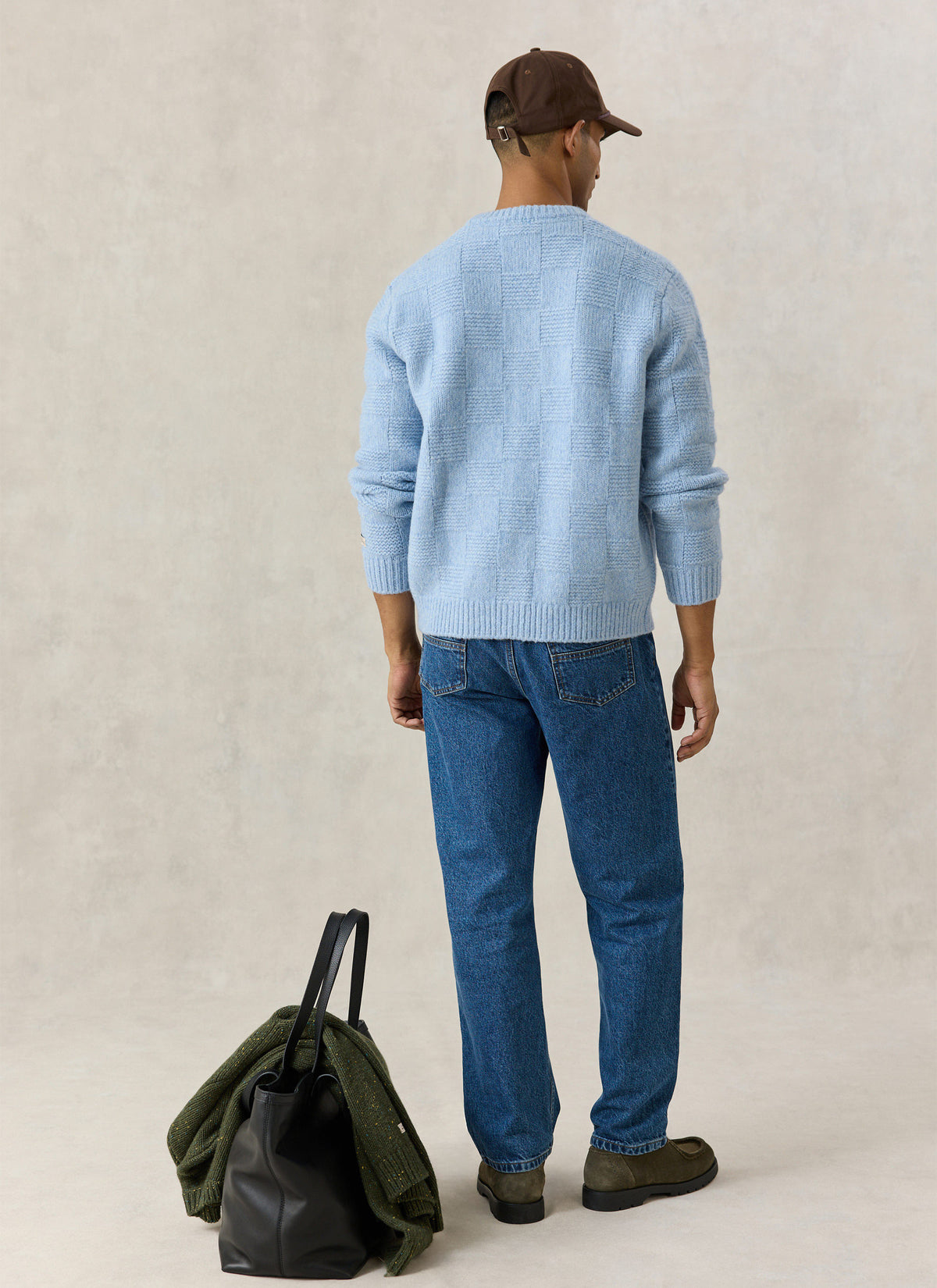 A model from the back wearing the Percival light blue alpaca wool knit cardigan, blue jeans, cap, and carrying a black bag.