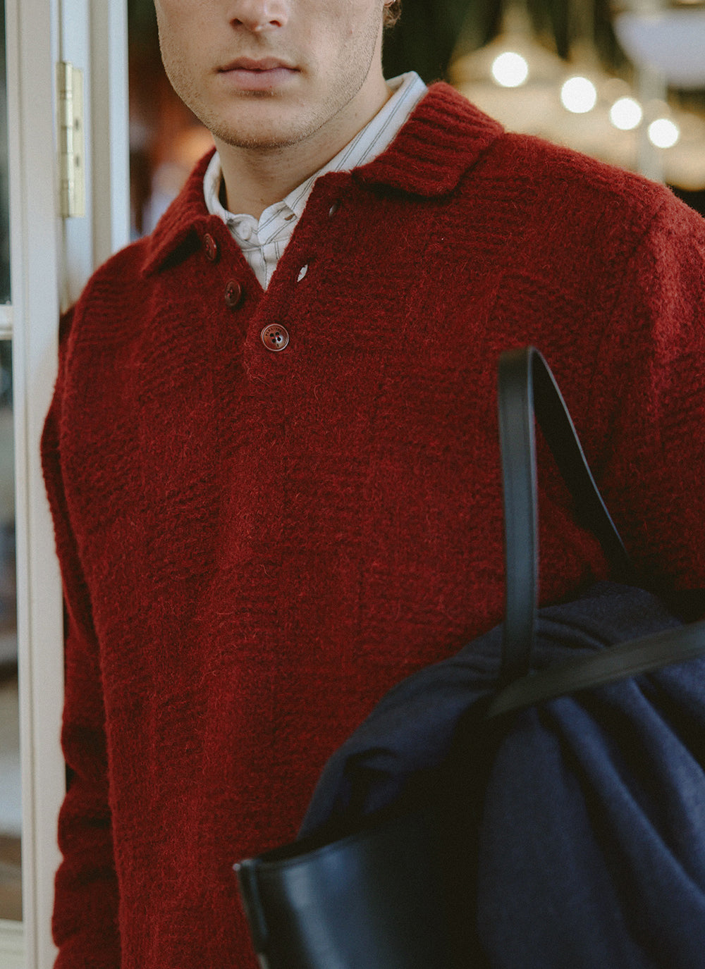Close-up of a model wearing the burgundy Percival Shugart knit jumper over a striped shirt, holding a dark bag.