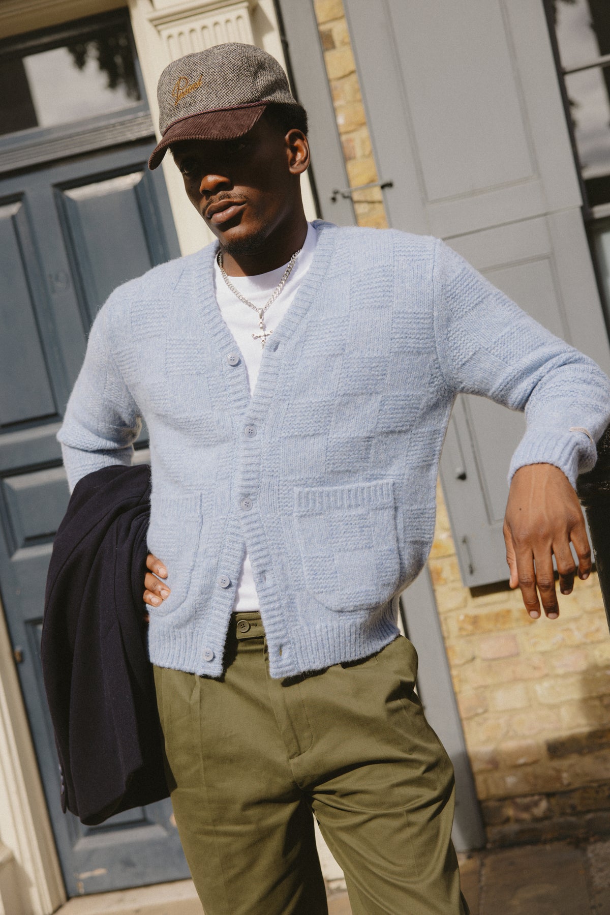 A model wearing the light blue Percival Shugart alpaca wool knit cardigan with green chinos, a cap and a jacket.