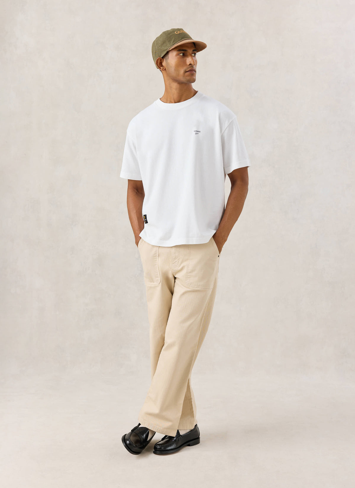 Model wearing the white Soloist oversized T-shirt from Guinness x Percival with beige trousers and a cap.
