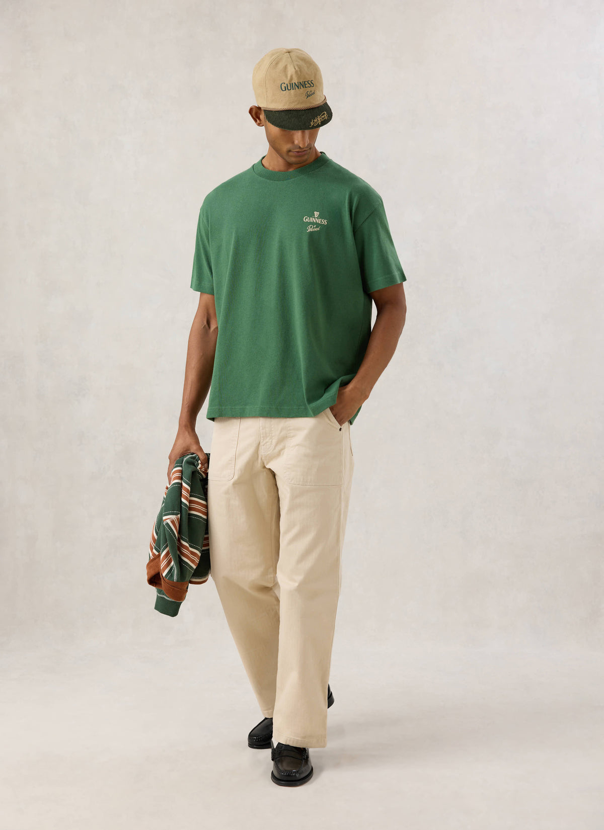 Model wearing the Guinness x Percival oversized T-shirt in forest green with chest print, beige trousers and cap.