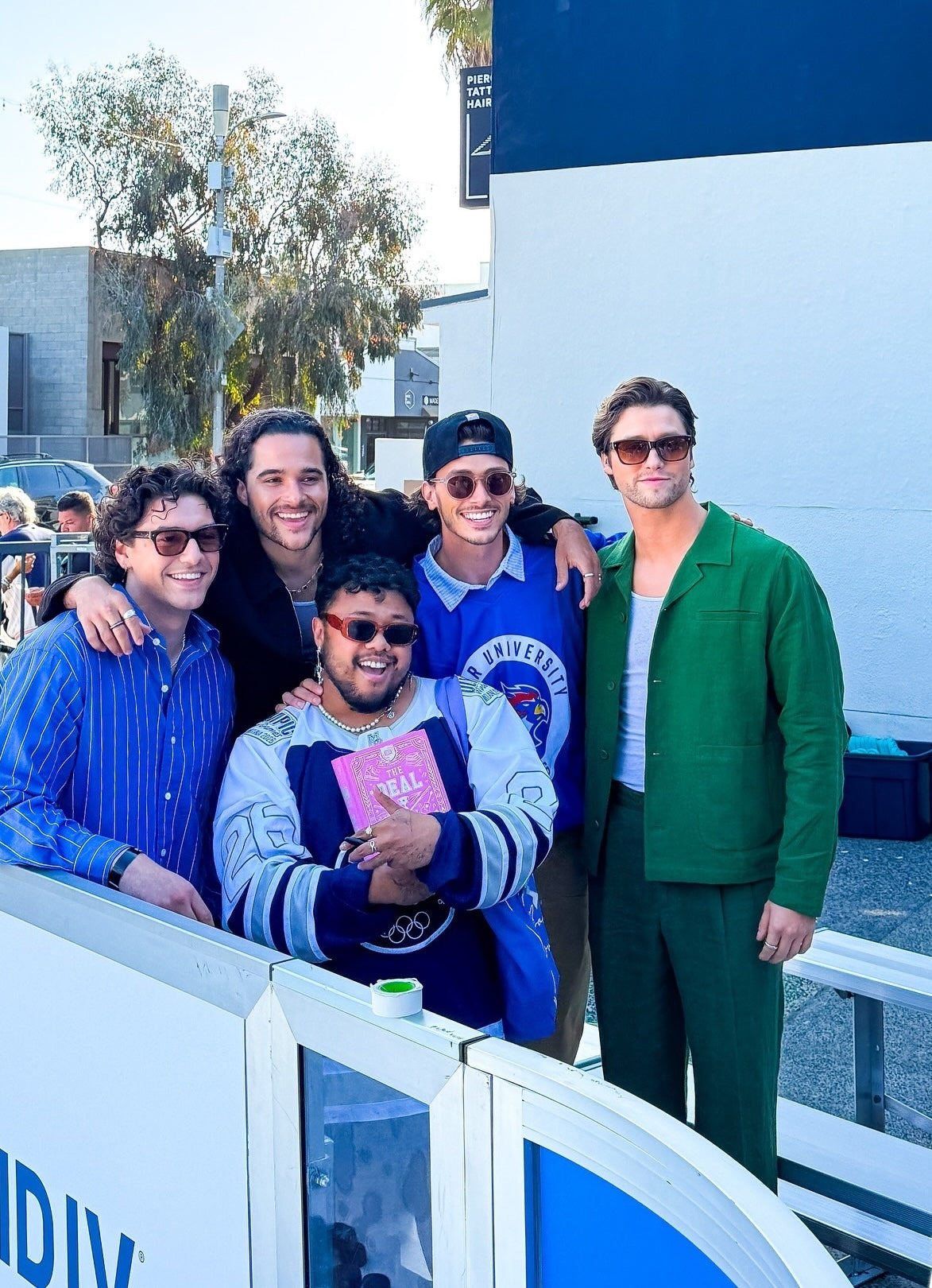 Actor Stephen Kalyn from Off Campus wearing the Percival boxy linen overshirt and matching linen trousers in forest green, standing with a group outdoors.