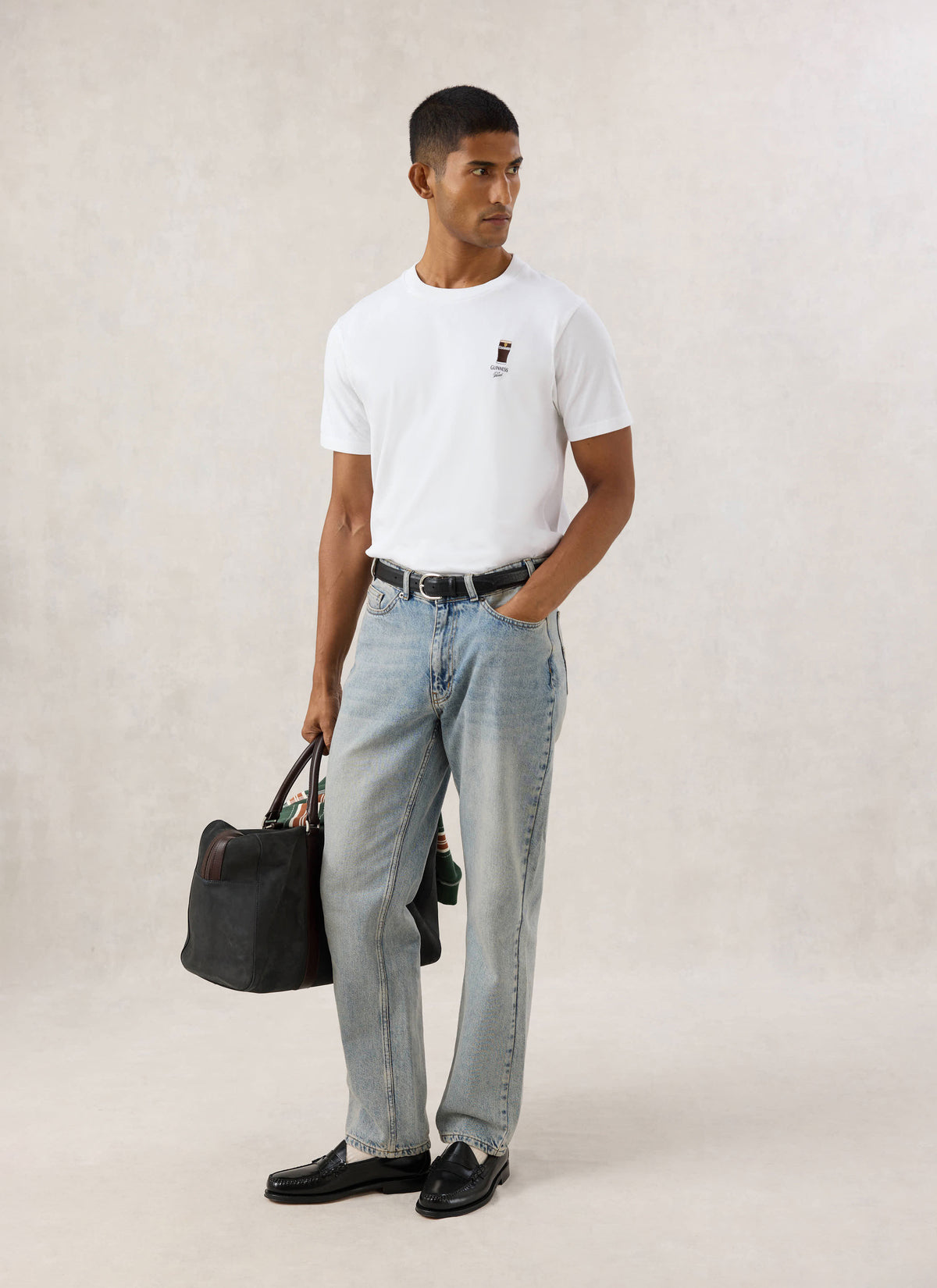 Model wearing the white Guinness x Percival stout embroidered T-shirt with light wash jeans and carrying a dark bag.