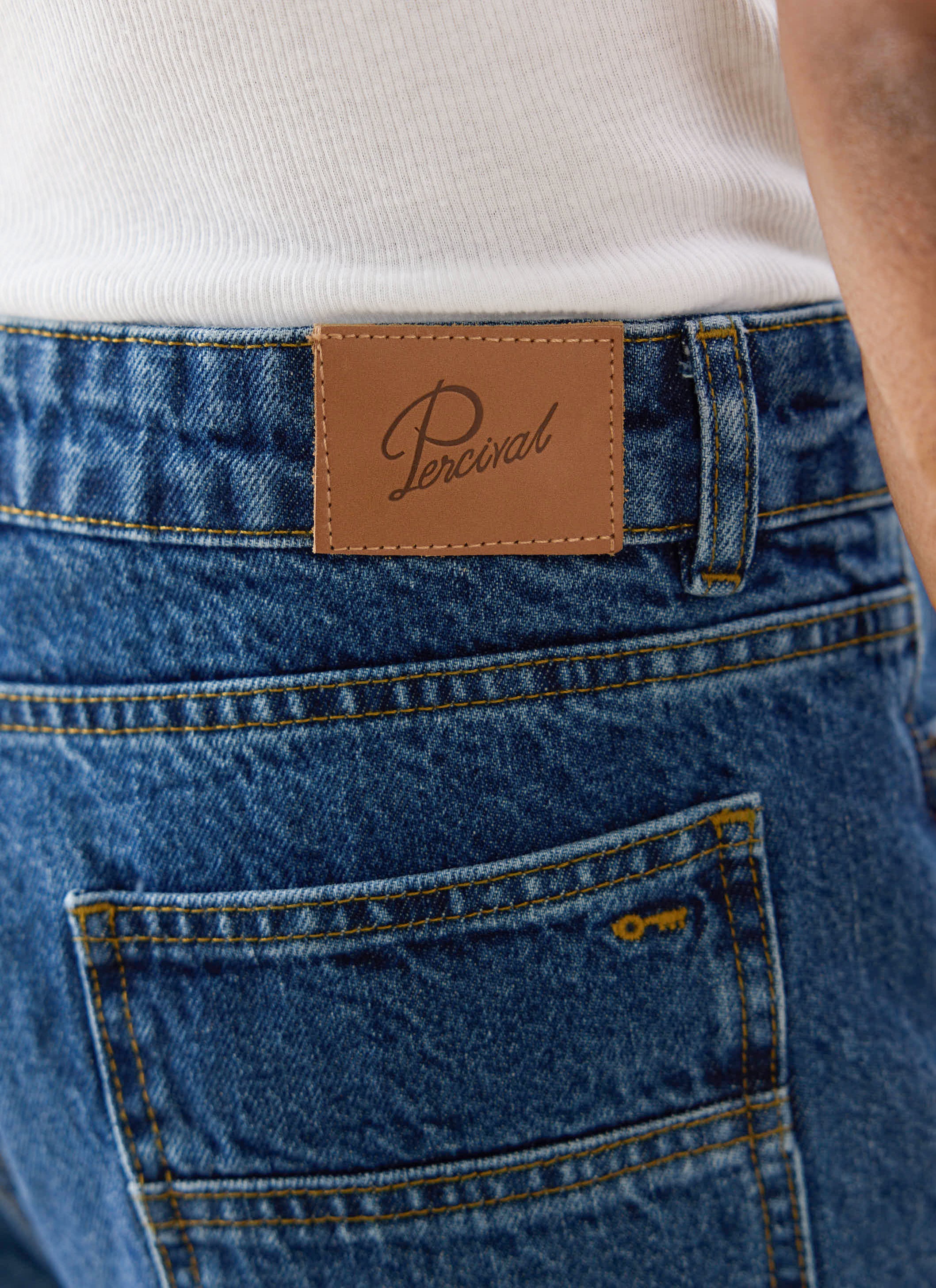 Close-up of Percival mid wash blue Heritage denim jeans, showing the leather branded patch and yellow embroidered key.
