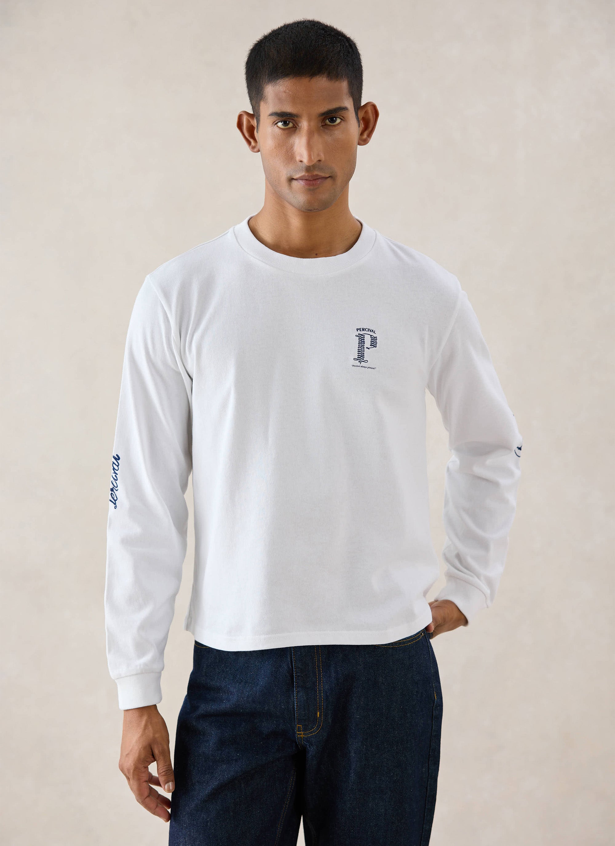 A model wears the white Percival Tucker Long Sleeve T-Shirt with graphic chest embroidery, styled with blue denim jeans.