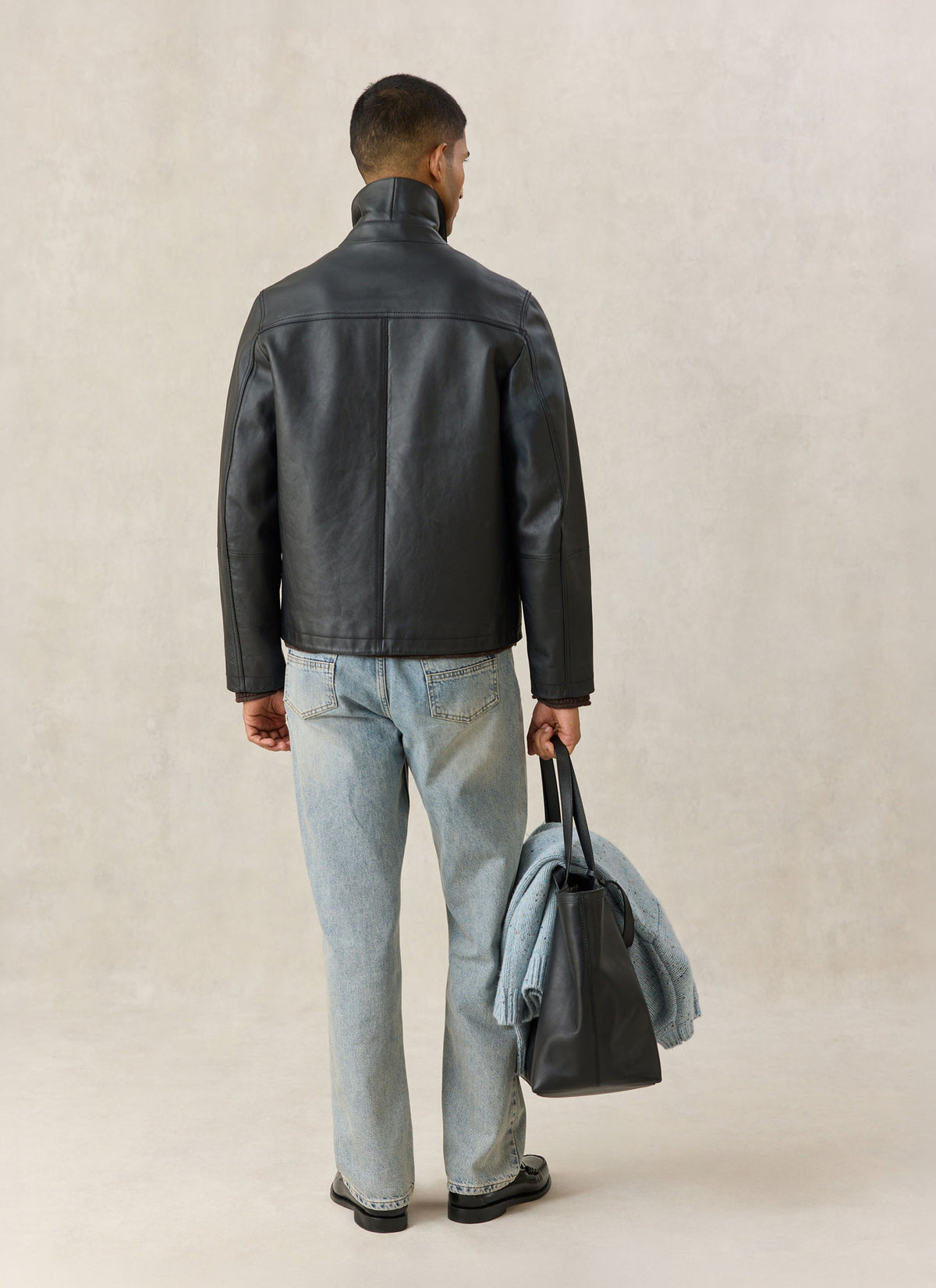 A model wearing the Percival black Turner leather jacket from the back, paired with light blue jeans and holding a dark bag.