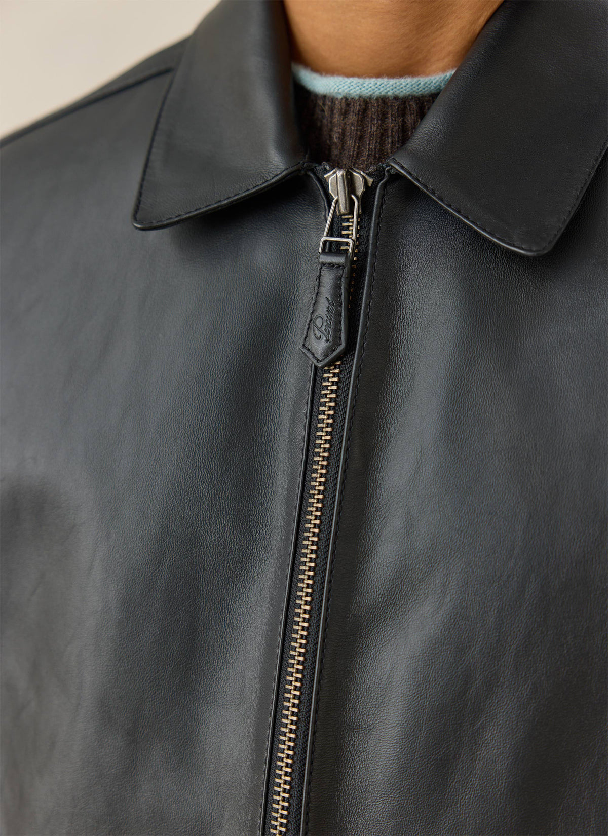 A close-up of the Percival Turner black leather zip jacket showing the collar, front metal zipper, and layered knitwear.