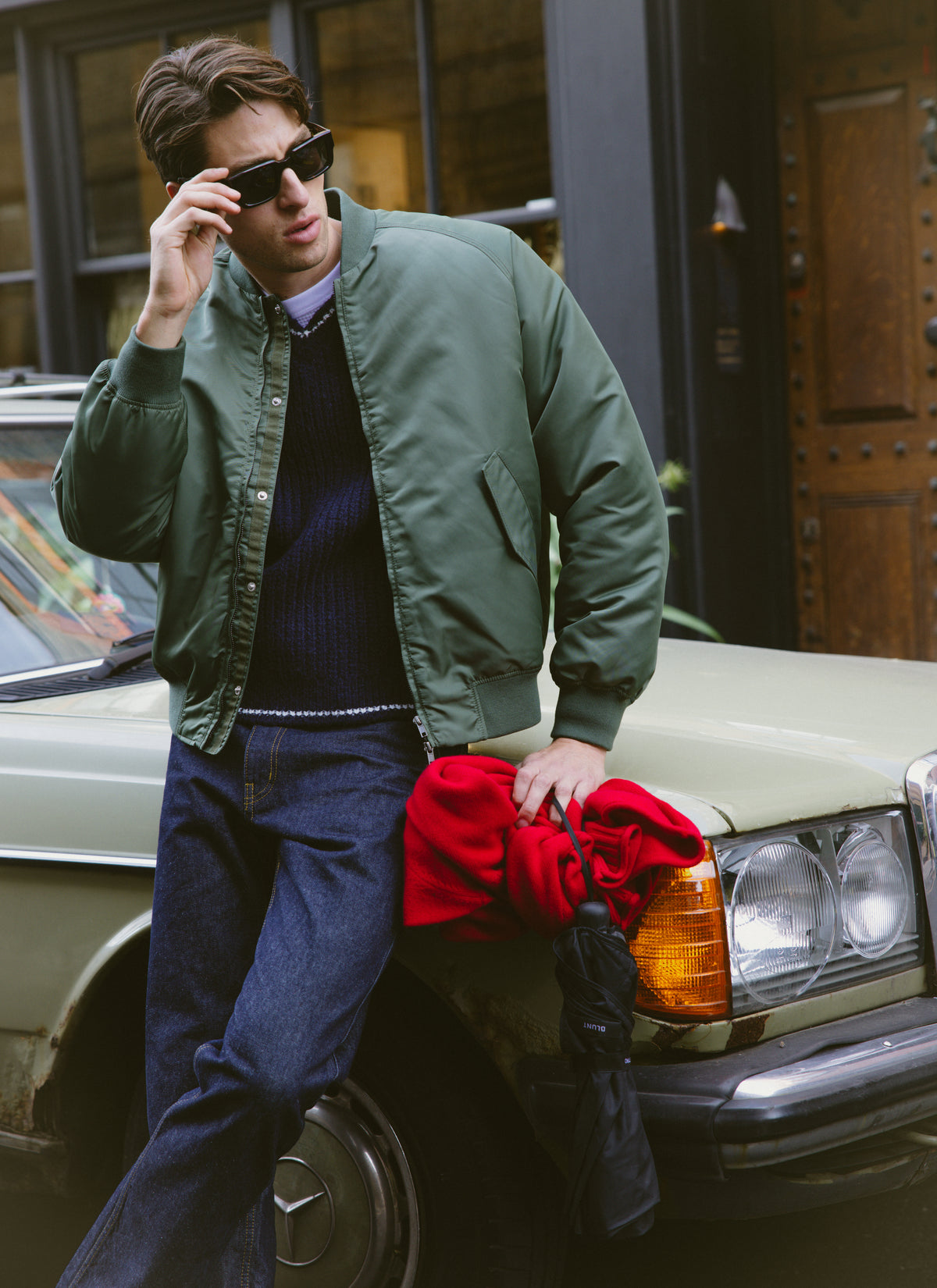 A model wearing a green bomber jacket and dark jeans leans against a classic car, holding the red Percival roll-neck jumper.