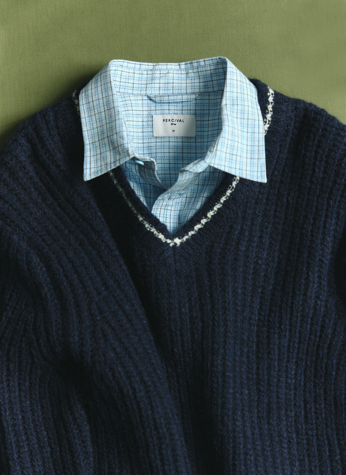 A close-up of the Percival navy alpaca wool V-neck jumper layered over a light blue checked shirt against a green background.