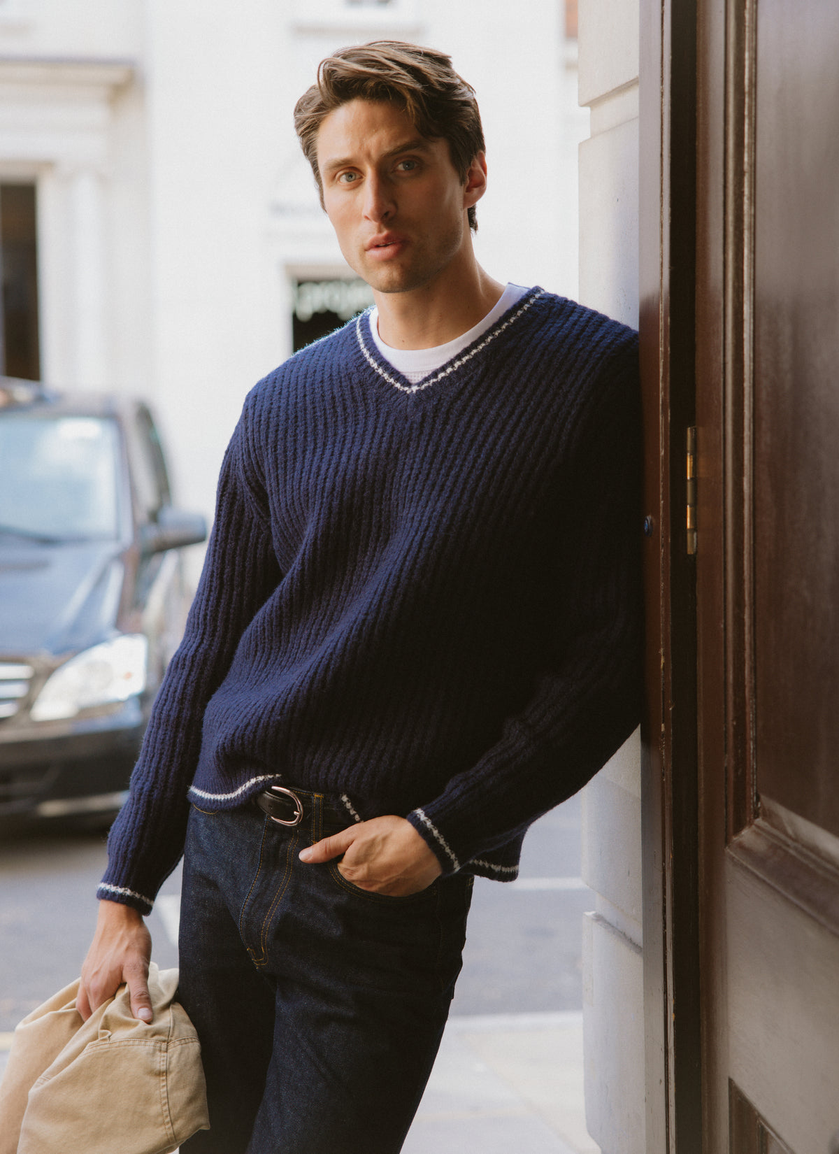 A model wearing the navy Percival Vincent V-neck jumper outdoors, leaning against a doorway.