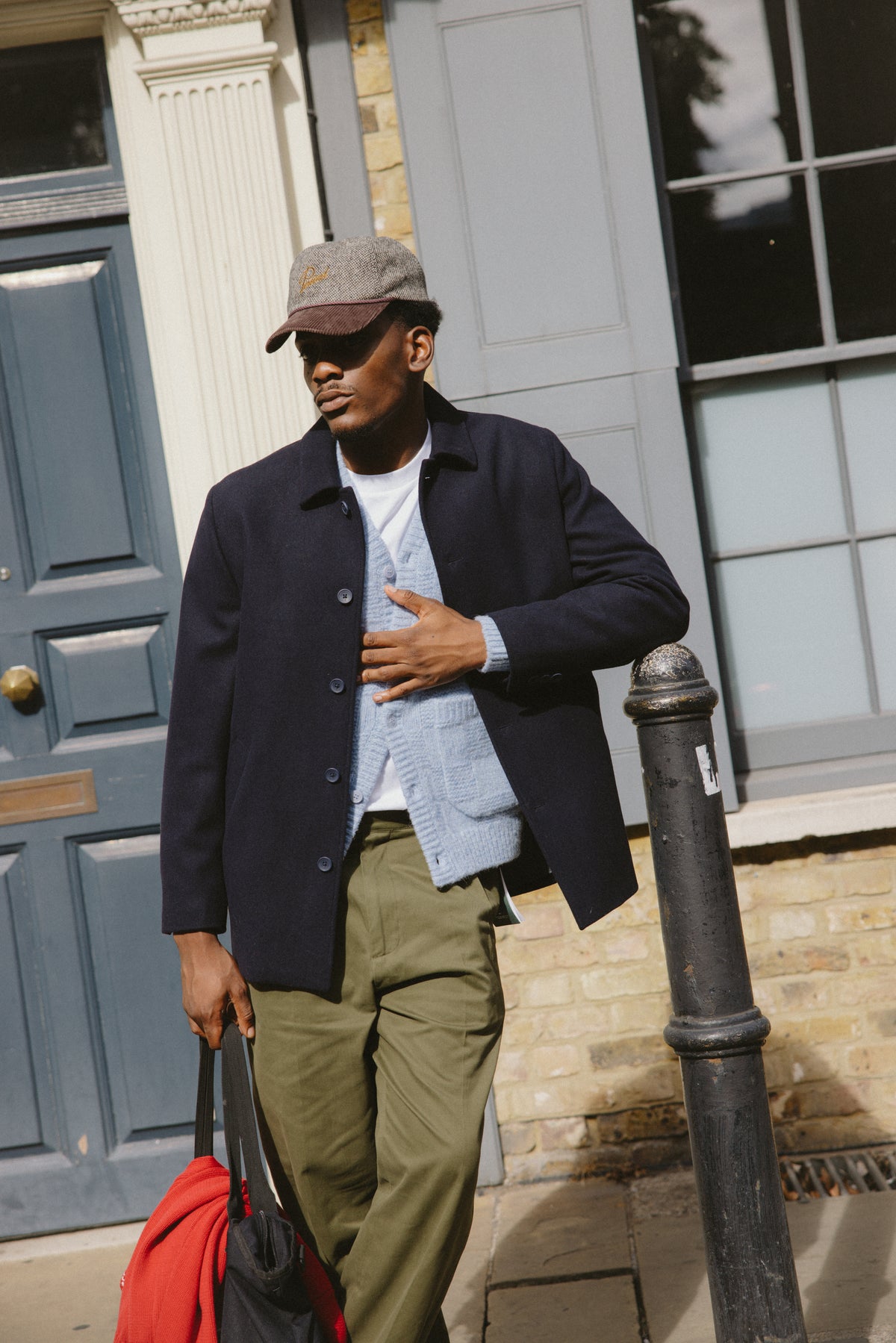 Model wearing the light blue alpaca wool Shugart cardigan under a dark jacket and green chinos on an outdoor street.