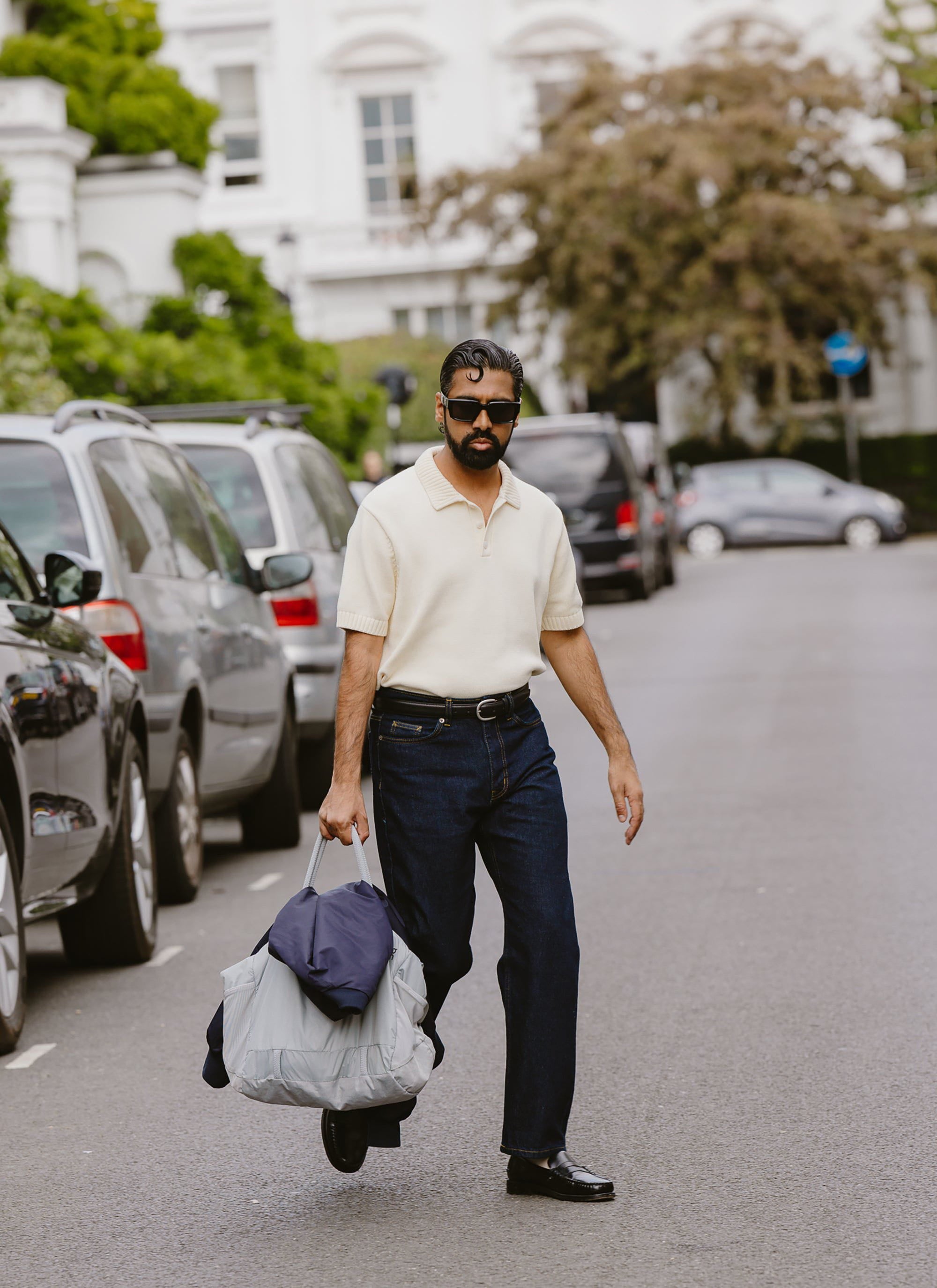 A model wears the ecru Percival Verso Knitted Polo with dark jeans and sunglasses, walking down a London street.