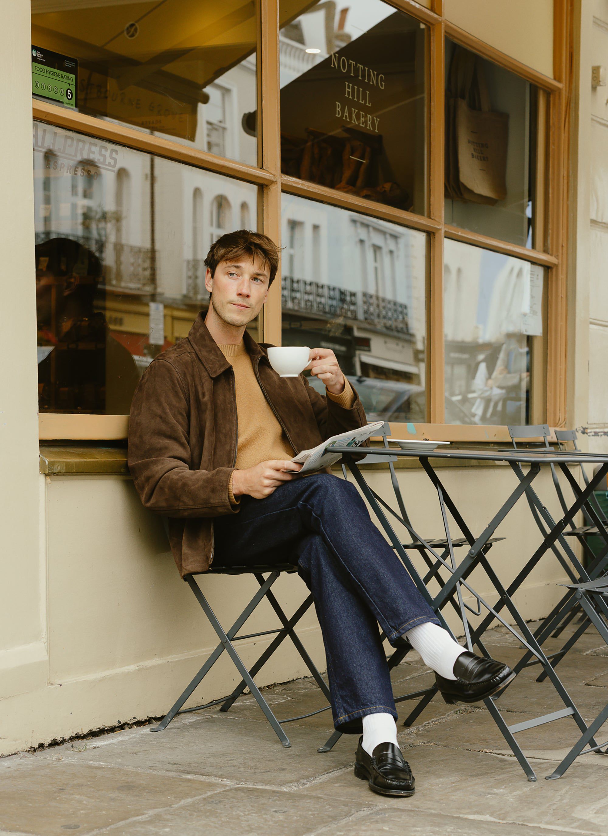 A man wearing the Percival brown Felted Wool Jumper layered under a jacket, sits outside a Notting Hill bakery with a coffee.