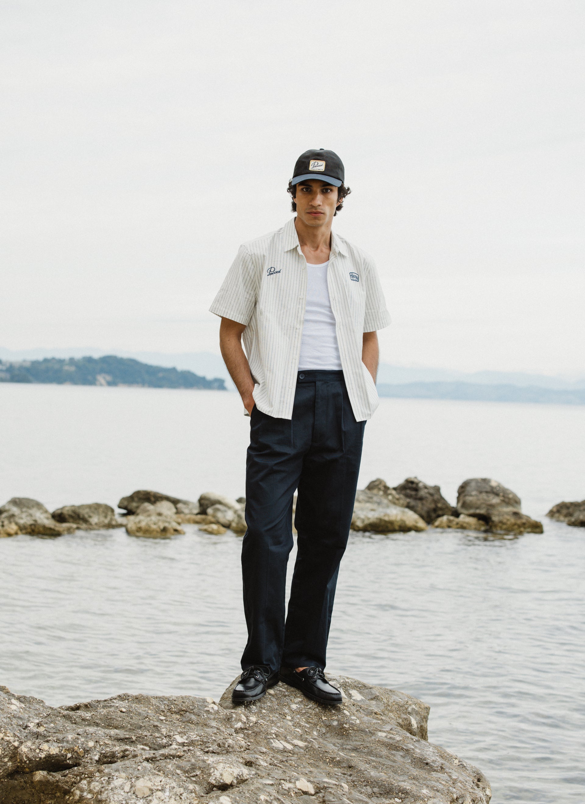 Model in the Percival Lucia cream stripe cotton embroidered shirt and navy Blake trousers on a rocky coastline.