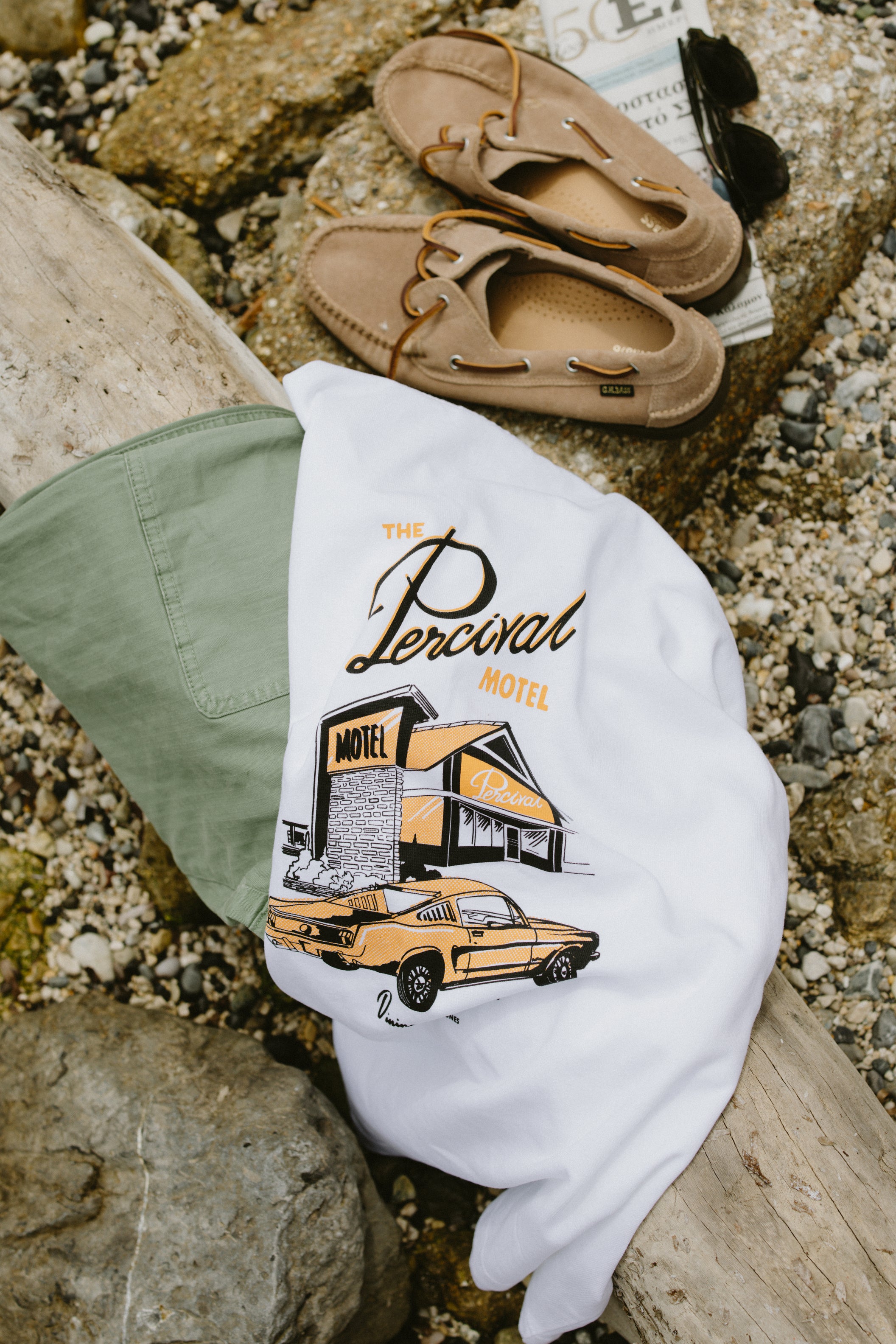 White Alistair oversized T-shirt by Percival featuring 'The Percival Motel' back graphic, styled on a pebbled beach.