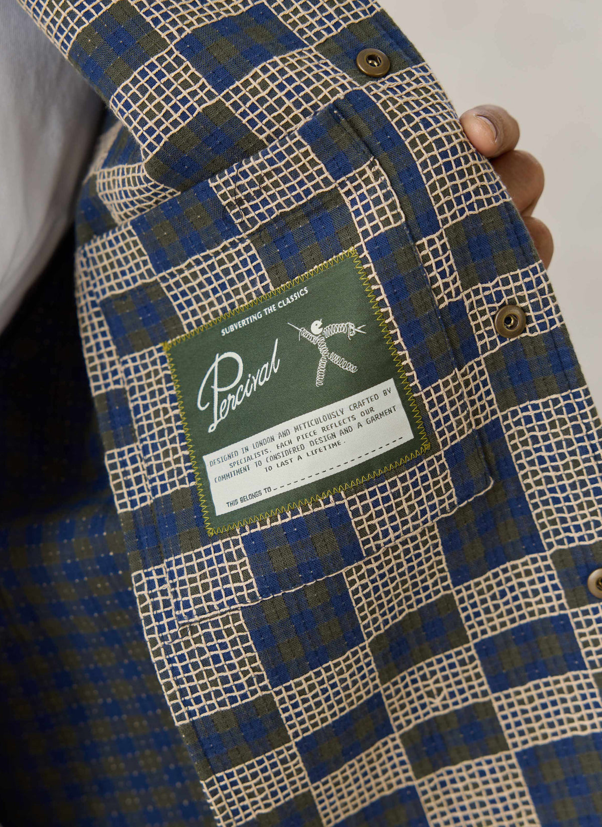 A close-up of the inside of the Percival Wallis Grid Check Overshirt showing the green brand label and cotton fabric.
