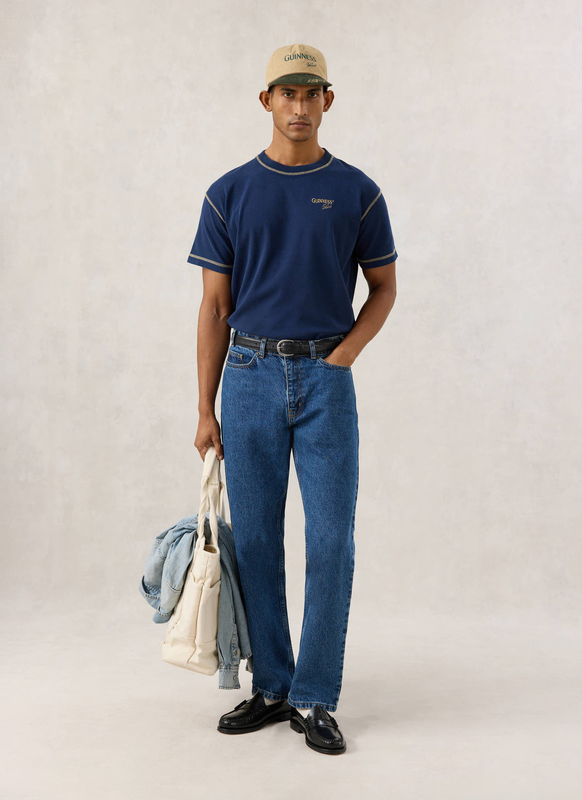 Model wearing the navy Guinness x Percival oversized T-shirt with contrast stitching, blue jeans, cap, and a tote bag.