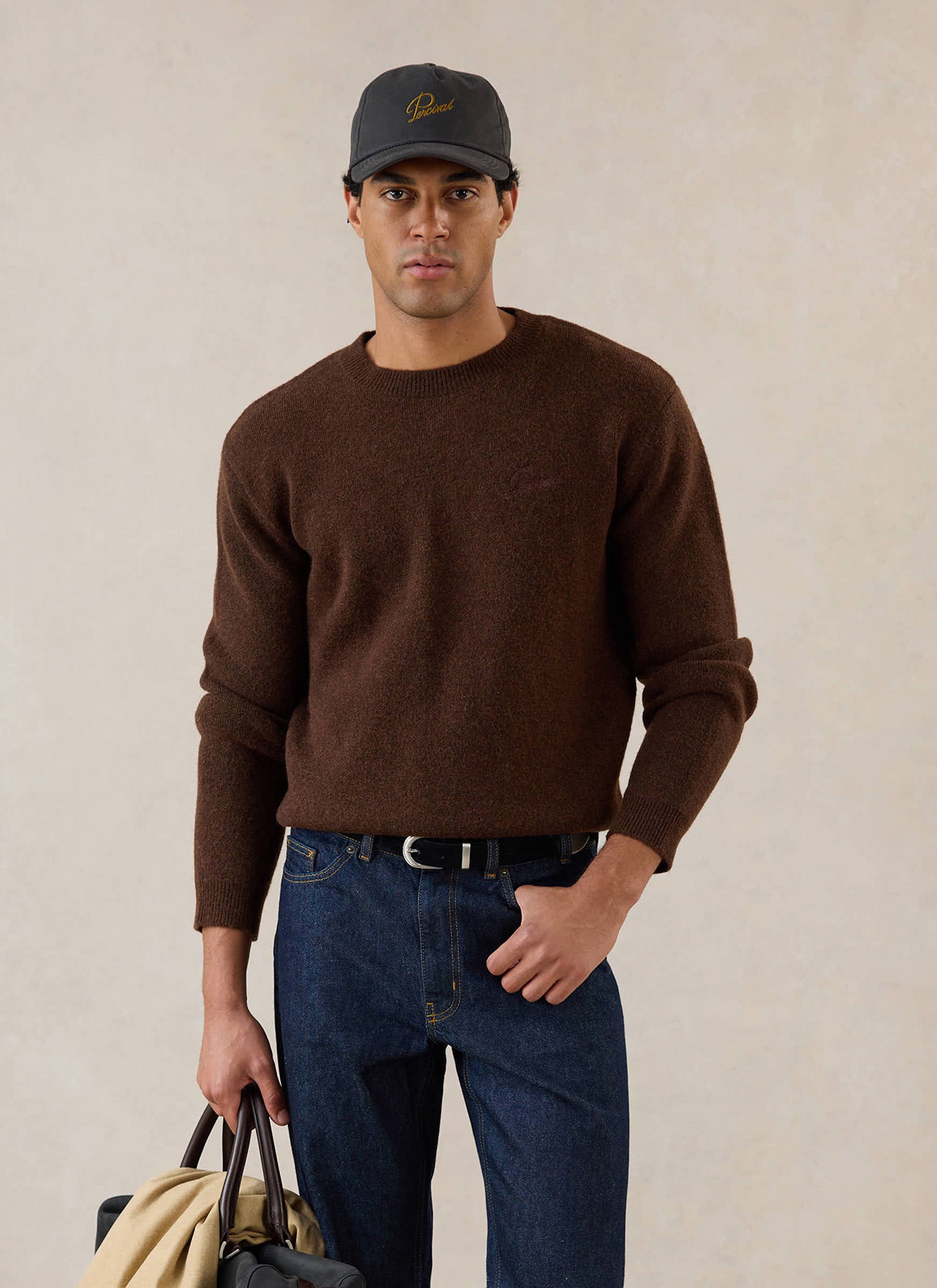 A model wears the espresso Percival White Glove boxy crew neck jumper, styled with dark blue denim jeans and a cap.