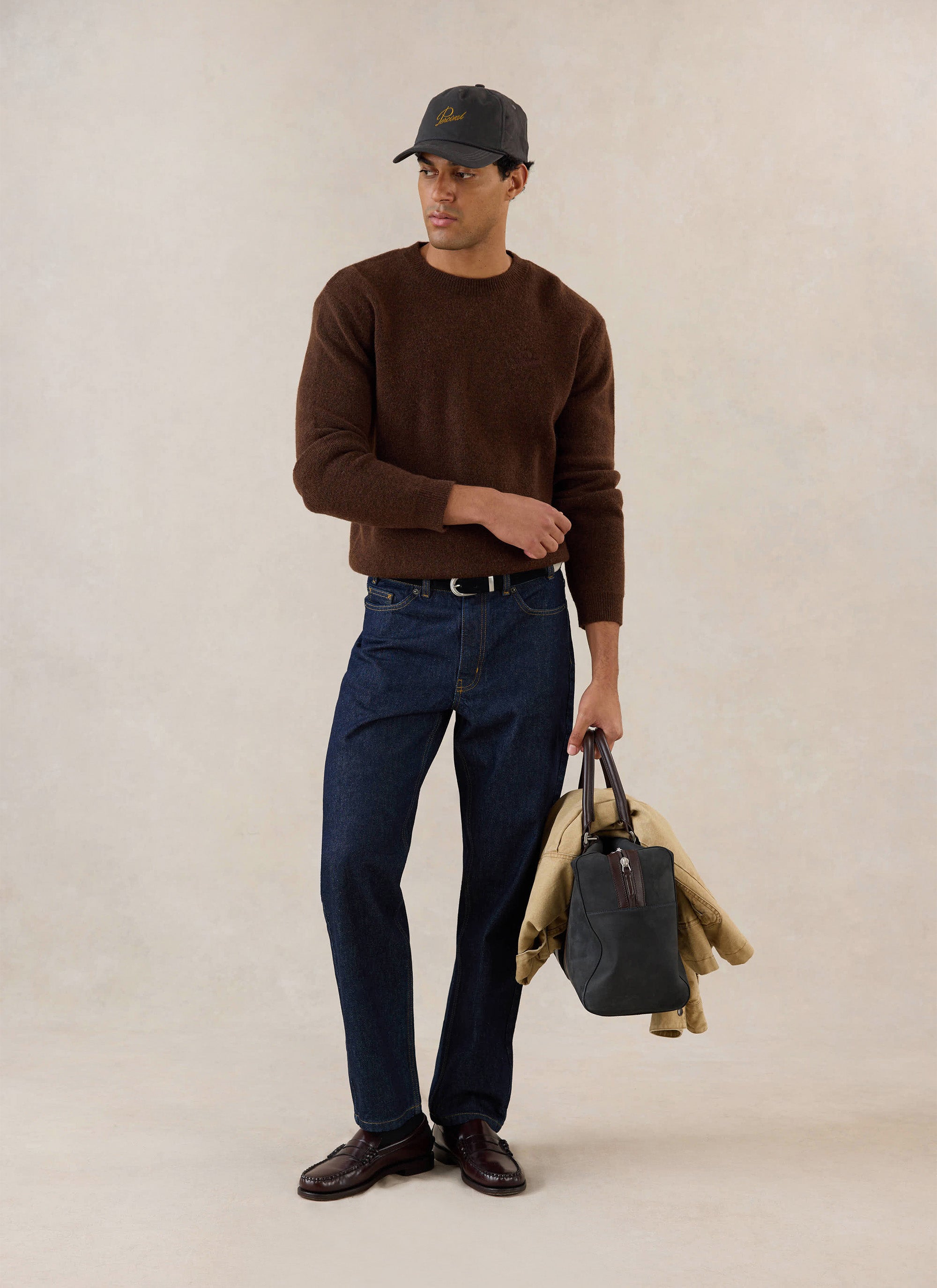 A model wears the espresso Percival mohair jumper with dark denim jeans, loafers, a cap, and a bag in a studio setting.