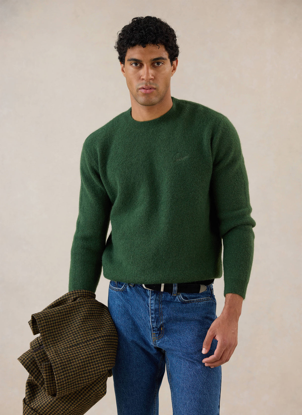 A model wears the forest green Percival mohair crew neck jumper with blue jeans, holding a houndstooth jacket.