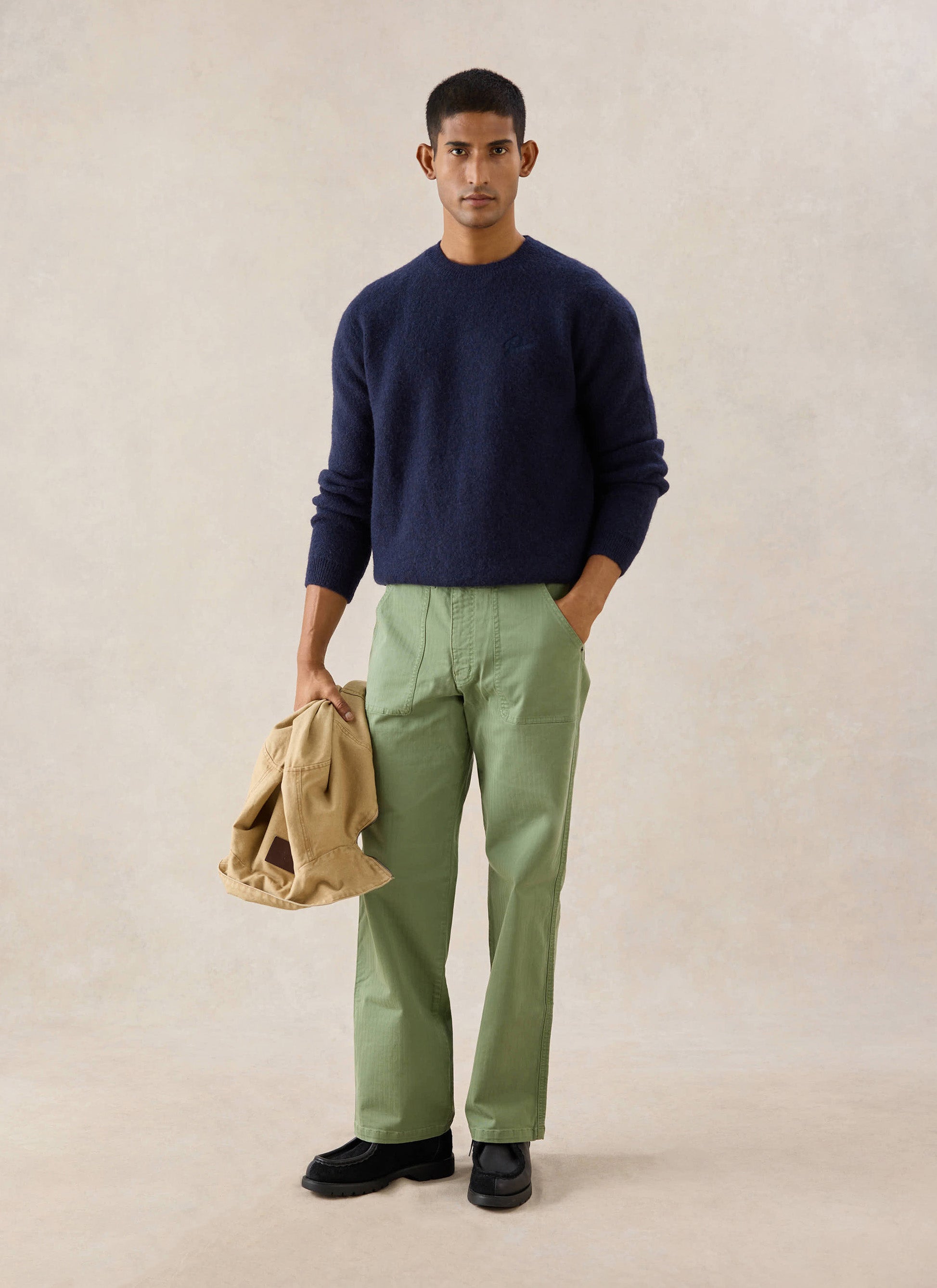 Model in navy Percival mohair crew neck jumper, green trousers, and a jacket in a studio setting.