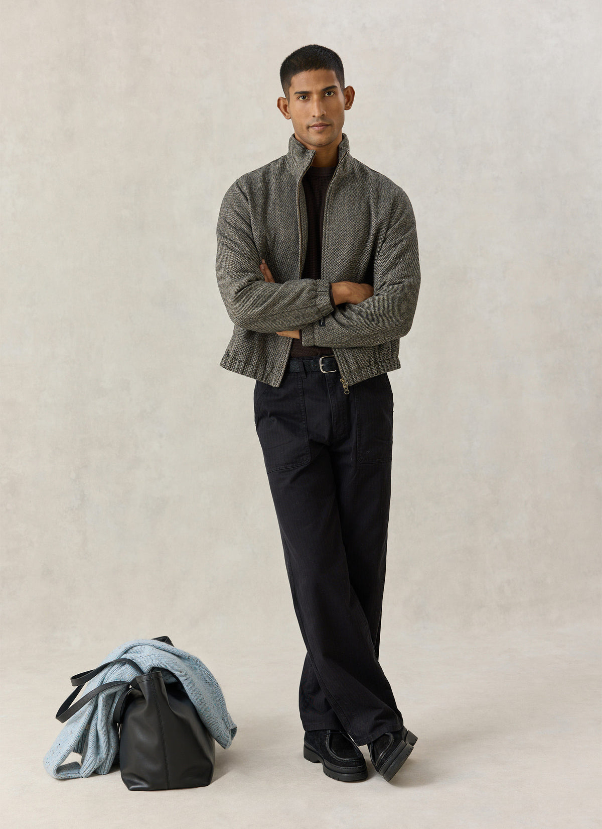A model wearing the Percival grey birdseye wool-blend Wilde Harrington jacket with dark trousers and a bag on the floor.