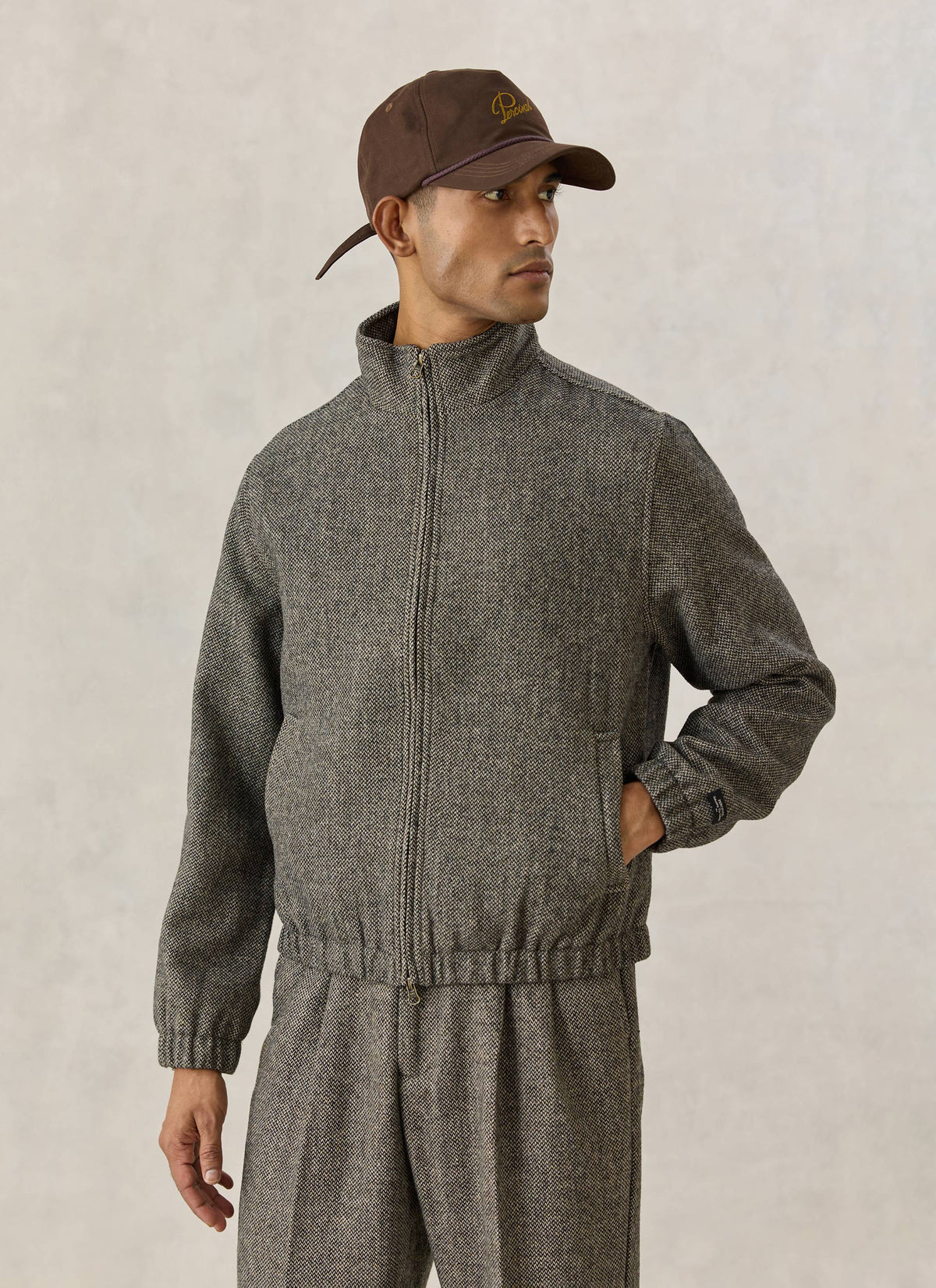 A model wearing the grey birdseye wool blend Percival Wilde Harrington jacket and matching trousers with a brown cap.