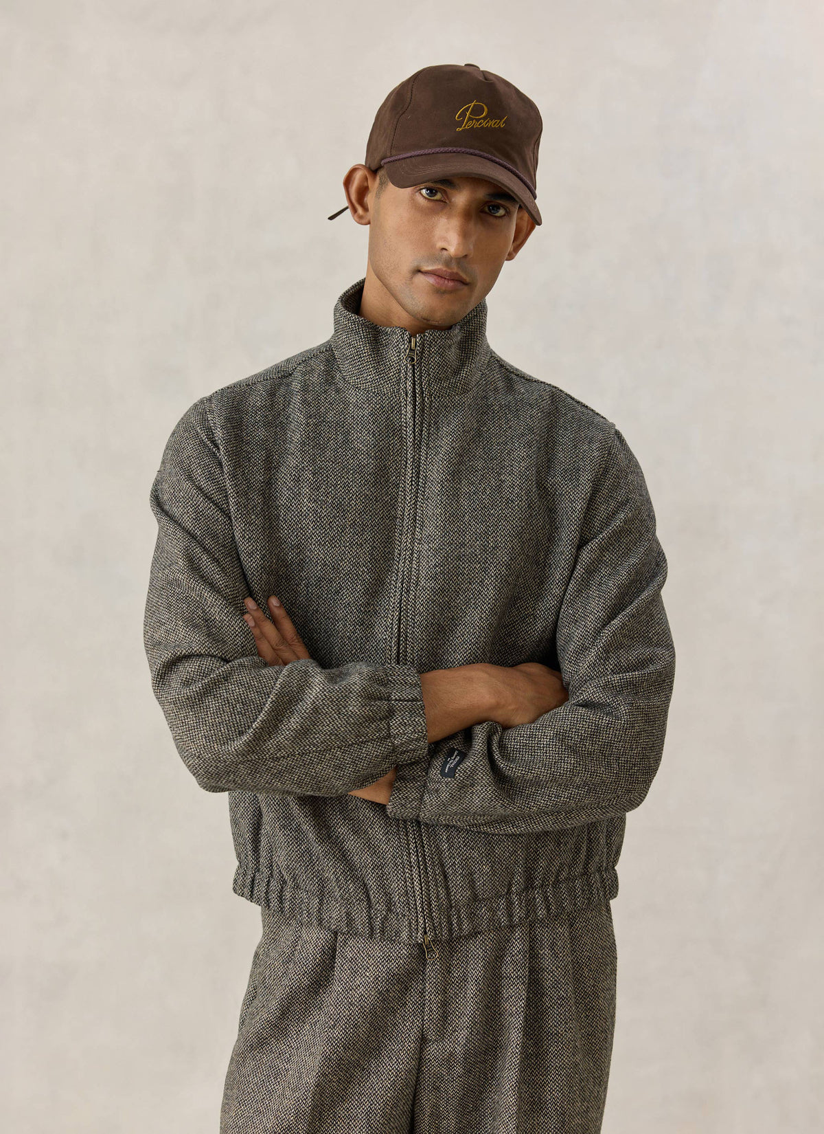 A model wearing the grey birdseye wool Percival Wilde Harrington jacket and trousers with a brown cap.
