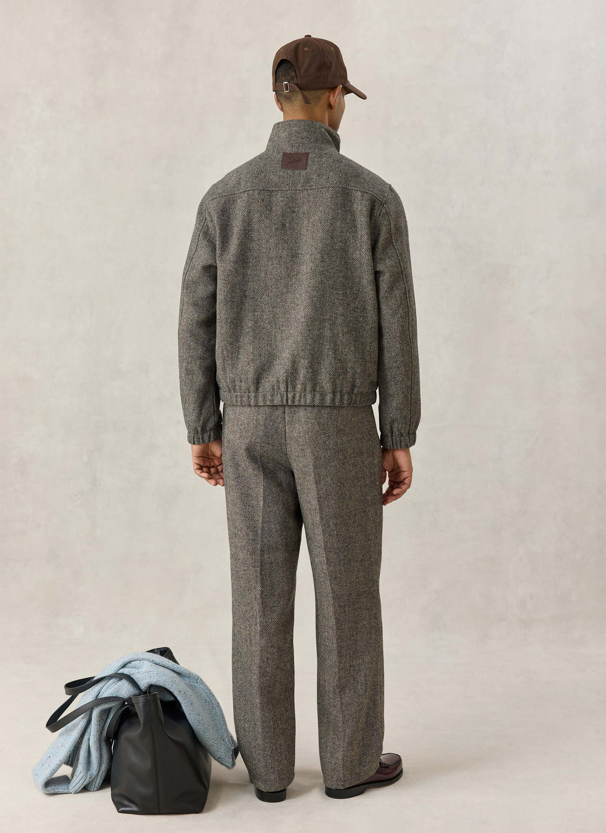 A model from the back wearing the grey birdseye wool-blend Wilde Harrington jacket, matching trousers, and a brown cap.