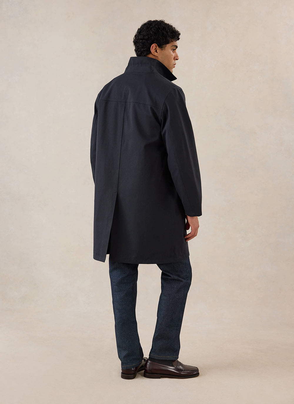 A rear view of a model wearing the black Wilson Long Trench Coat from Percival, styled with dark trousers and loafers.
