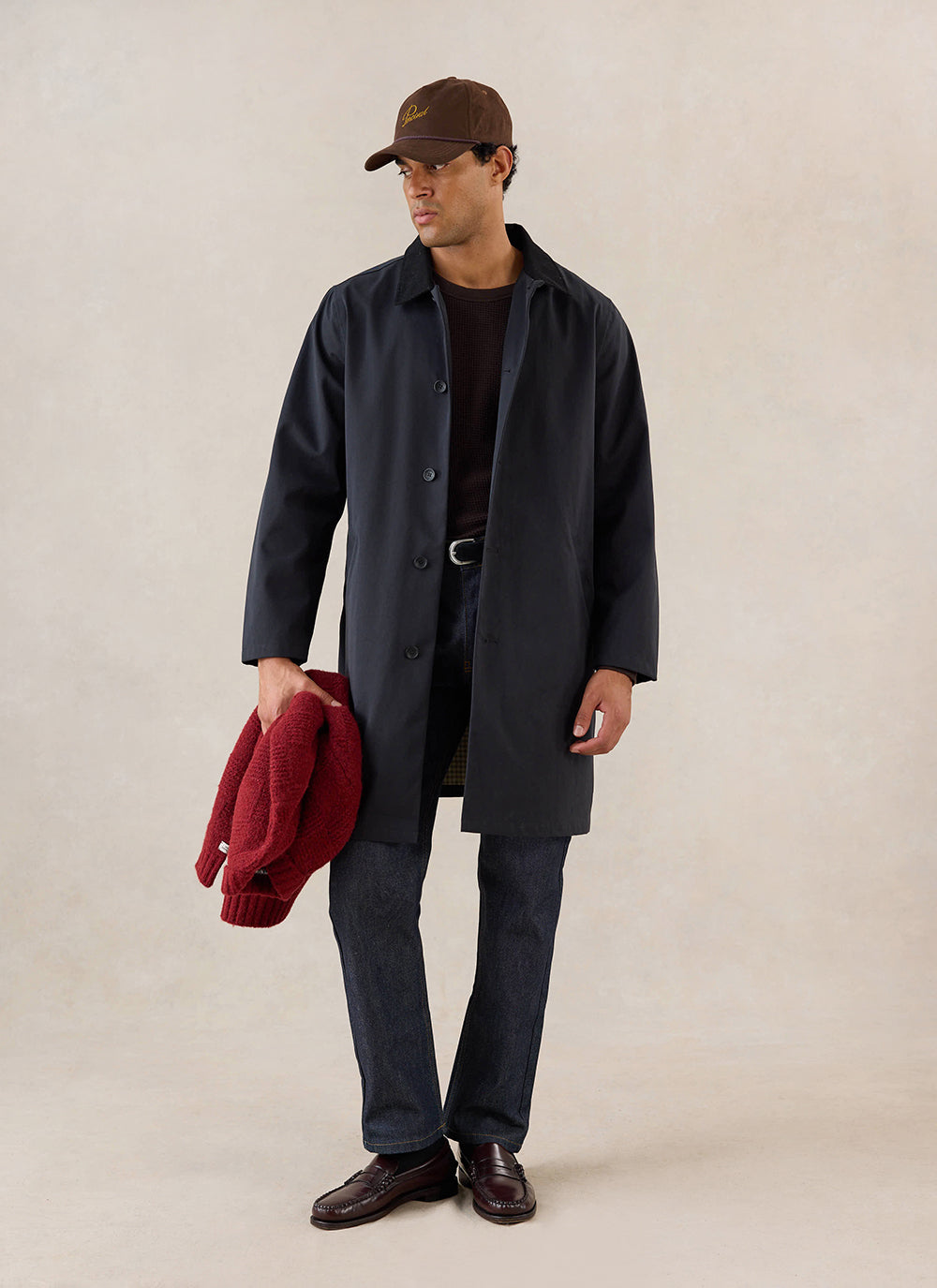A model wears the black Percival Wilson Long Trench Coat, styled with a brown cap, dark jeans, and loafers.