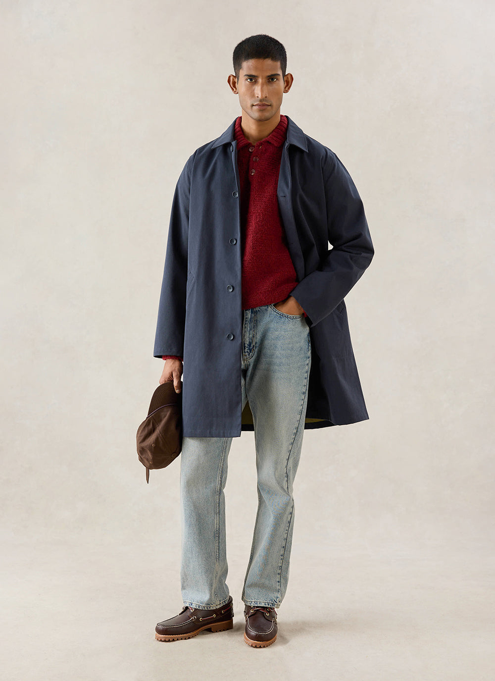 A model wears the midnight Wilson Long Trench Coat from Percival, styled with a red knit polo and light wash jeans.