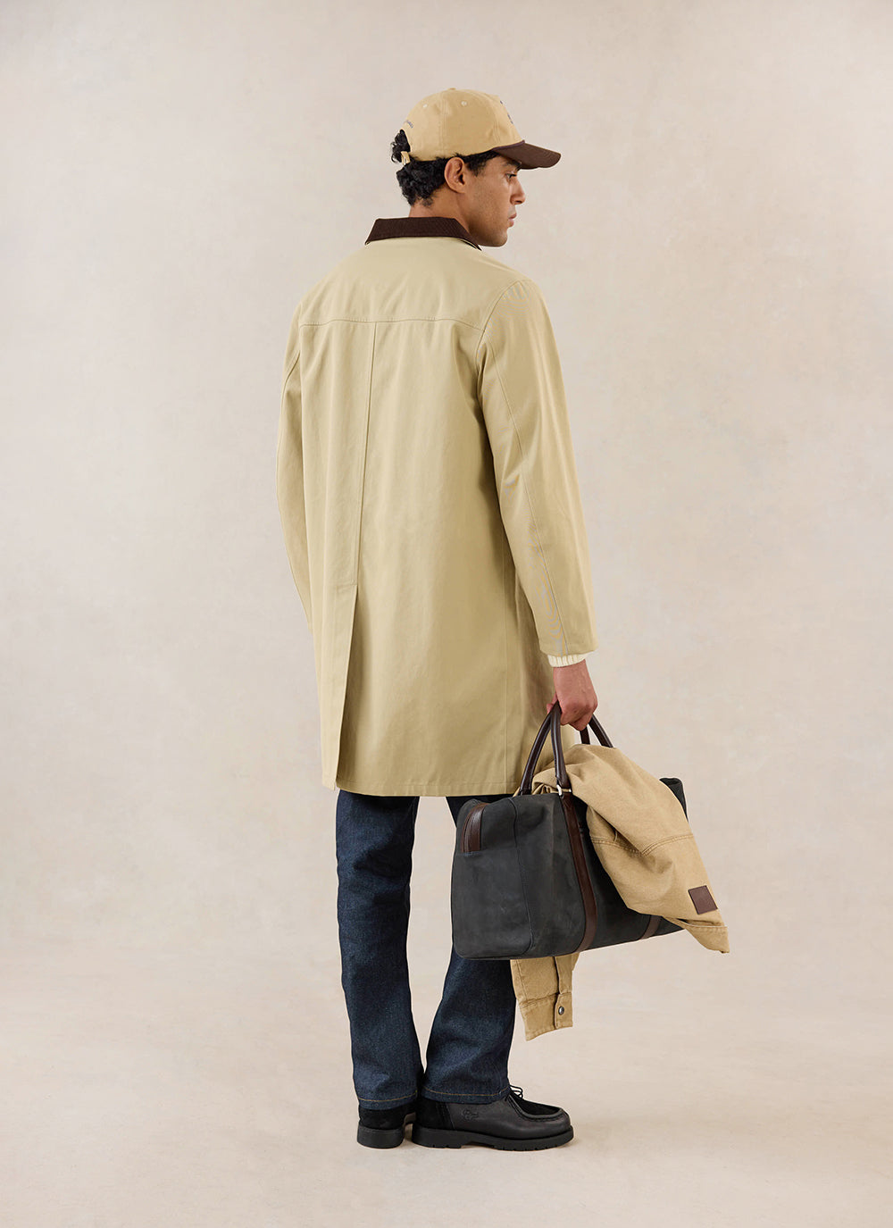 A model wears the tan Percival Wilson Long Trench Coat from behind, holding a dark holdall bag and cap in a studio.