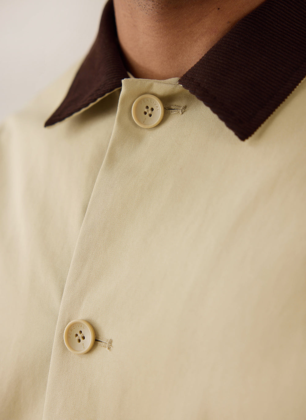 Close-up of the tan Percival Wilson trench coat, showing the contrasting brown corduroy collar and branded buttons.