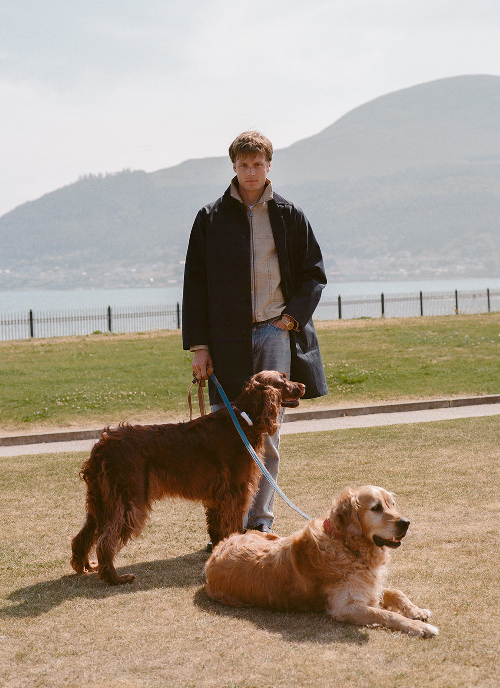 A model in a Percival waterproof trench coat and jacket with two dogs outdoors by the water.
