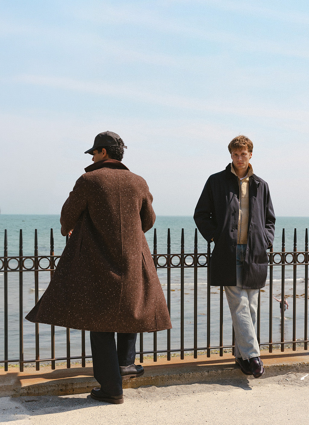 Two models wearing Percival outerwear by the seaside; one in a brown fleck overcoat, the other in a navy trench coat.